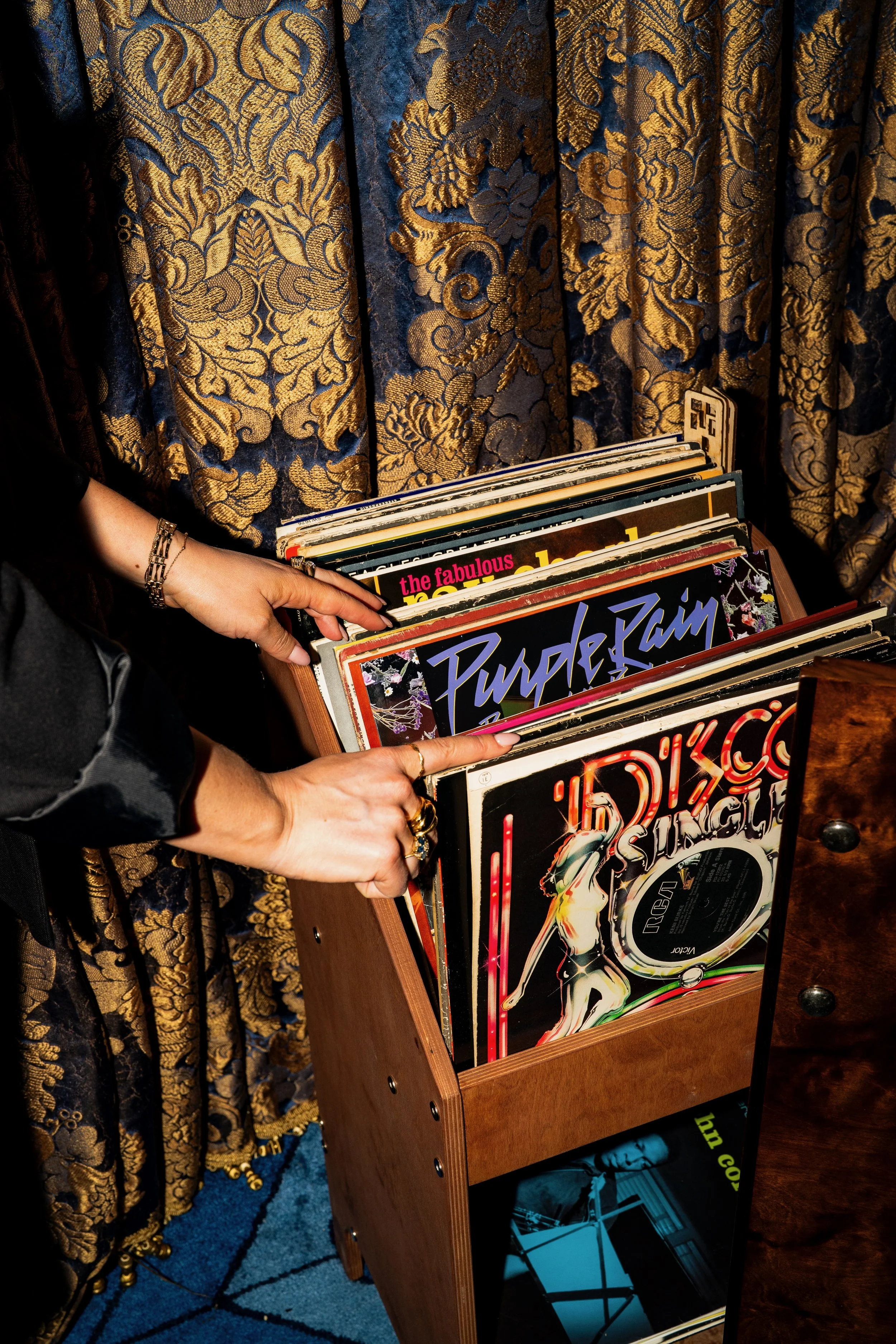 Close-up of a person's hand holding a vinyl record album in a wooden stand, with a background of ornate curtain fabric in gold and navy and a small part of a blue carpet.