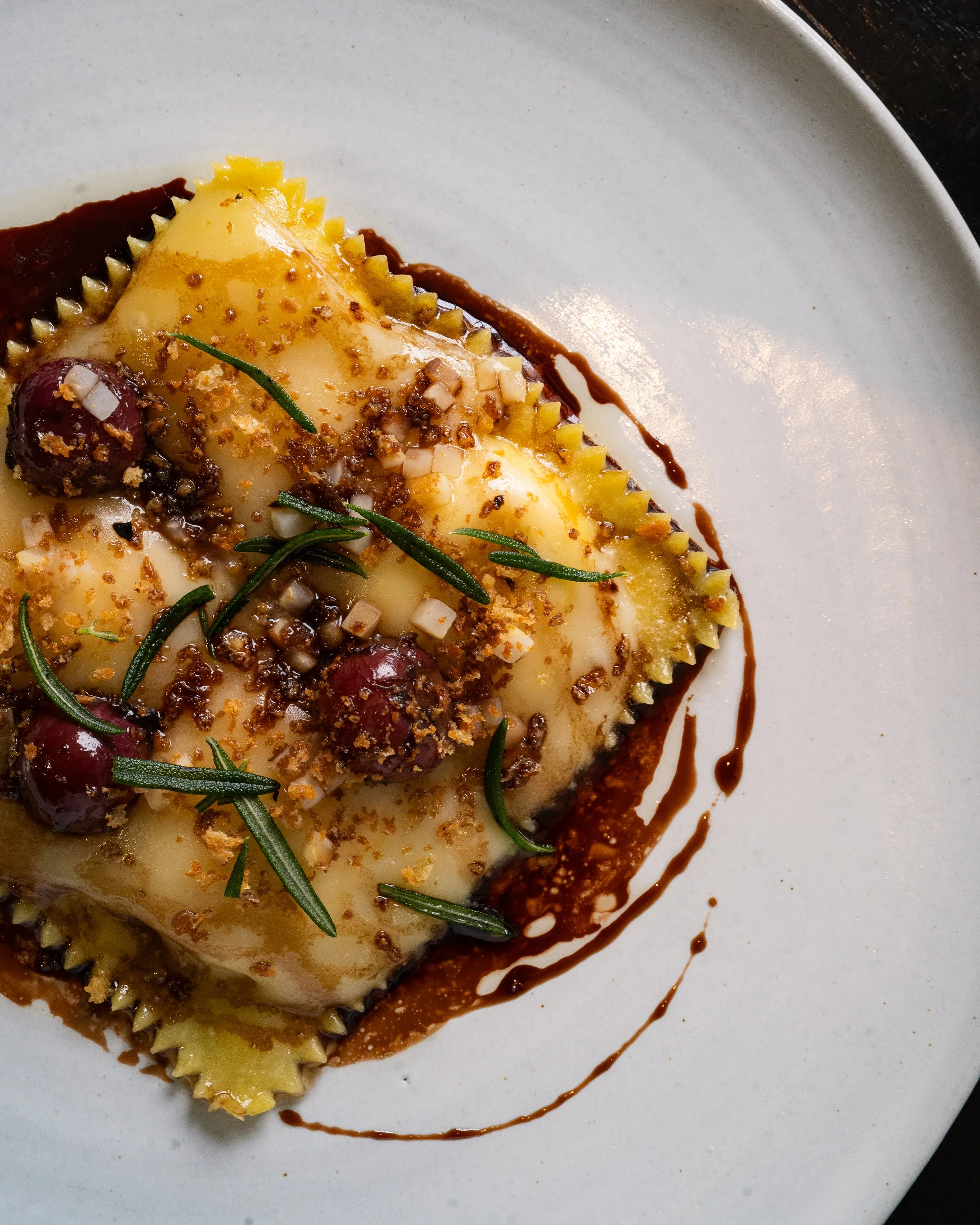 A plated dish featuring three ravioli pasta pieces topped with herbs, nuts, and a small amount of sauce, with a drizzle of sauce around on a white plate.