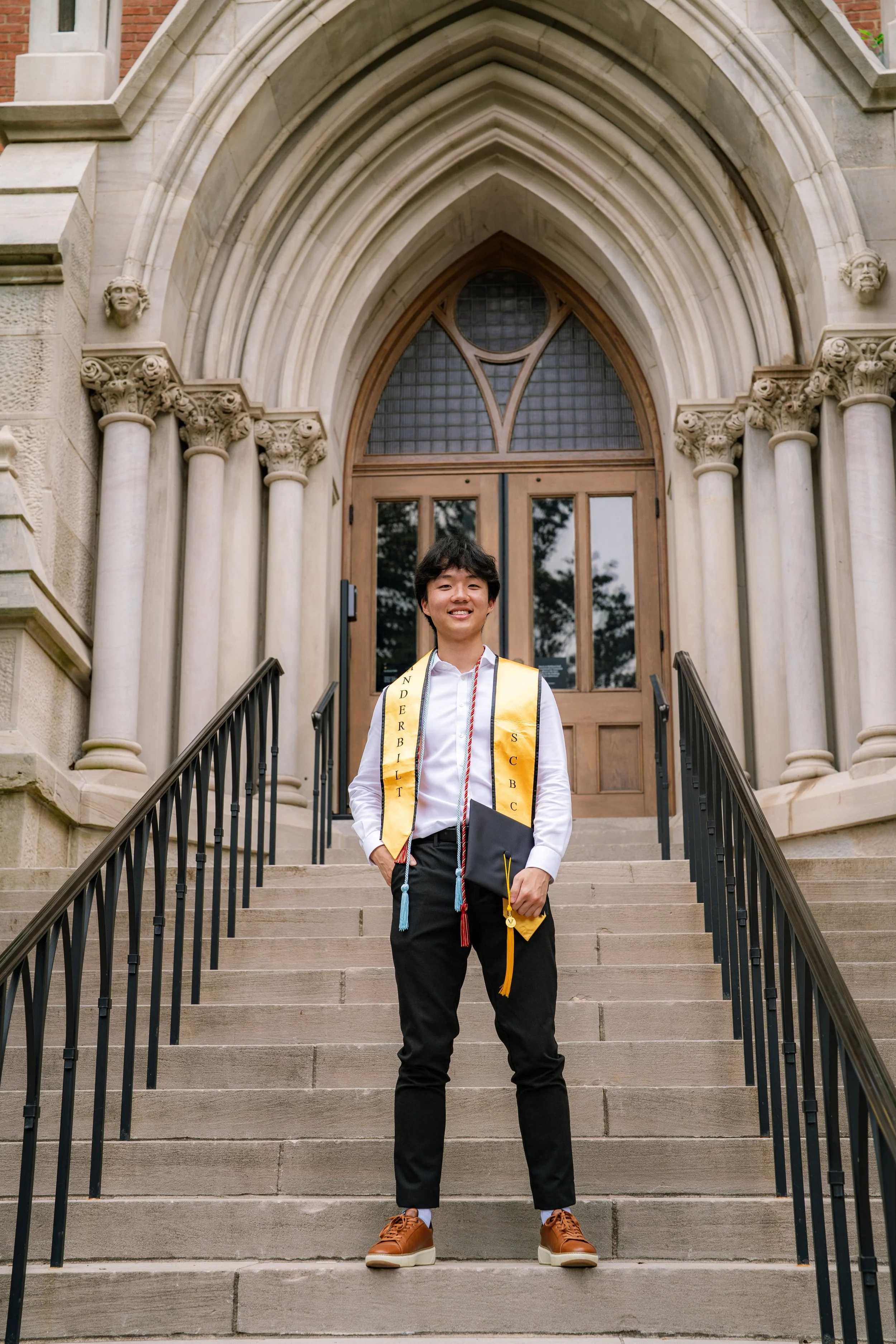 A young man in graduation attire stands on the steps of a historic building with archways and columns, smiling at the camera.