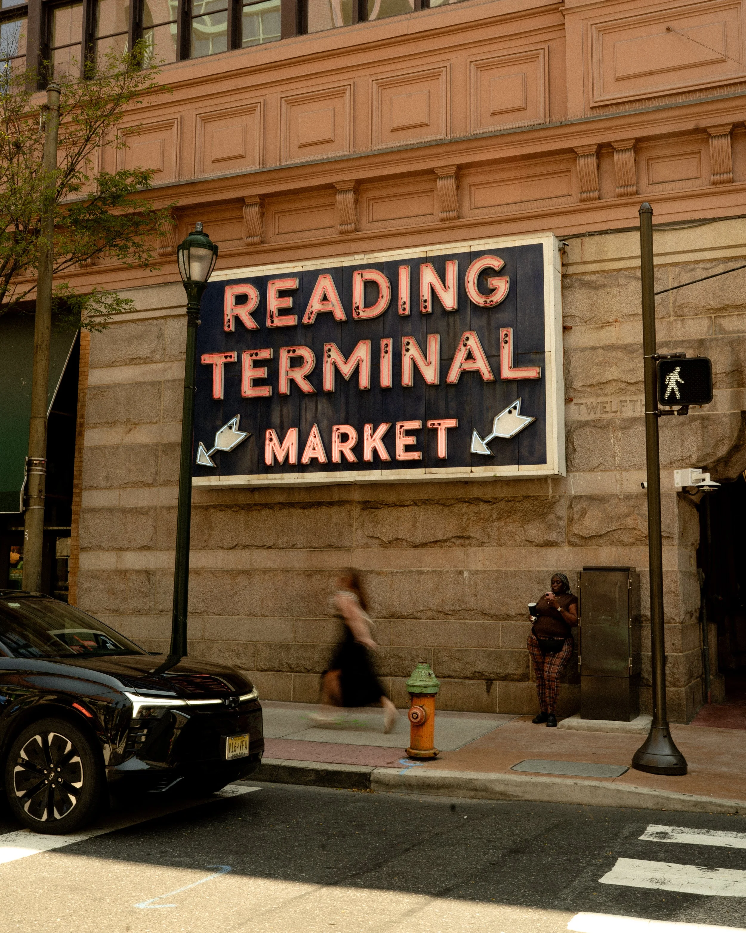The image shows the exterior of a building with a neon sign that reads 'Reading Terminal Market,' topped with decorative architectural molding. There are pedestrians on the sidewalk, a person with dark skin, glasses, and plaid pants standing near a b