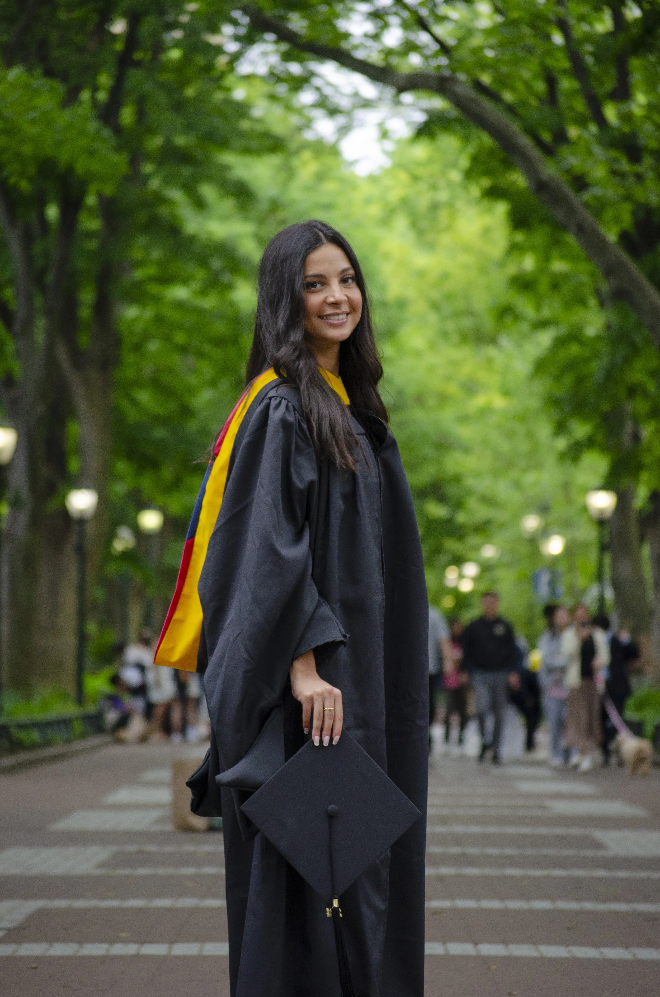 A young woman in a graduation cap and gown holding a diploma in a park-like setting with green trees and people in the background.