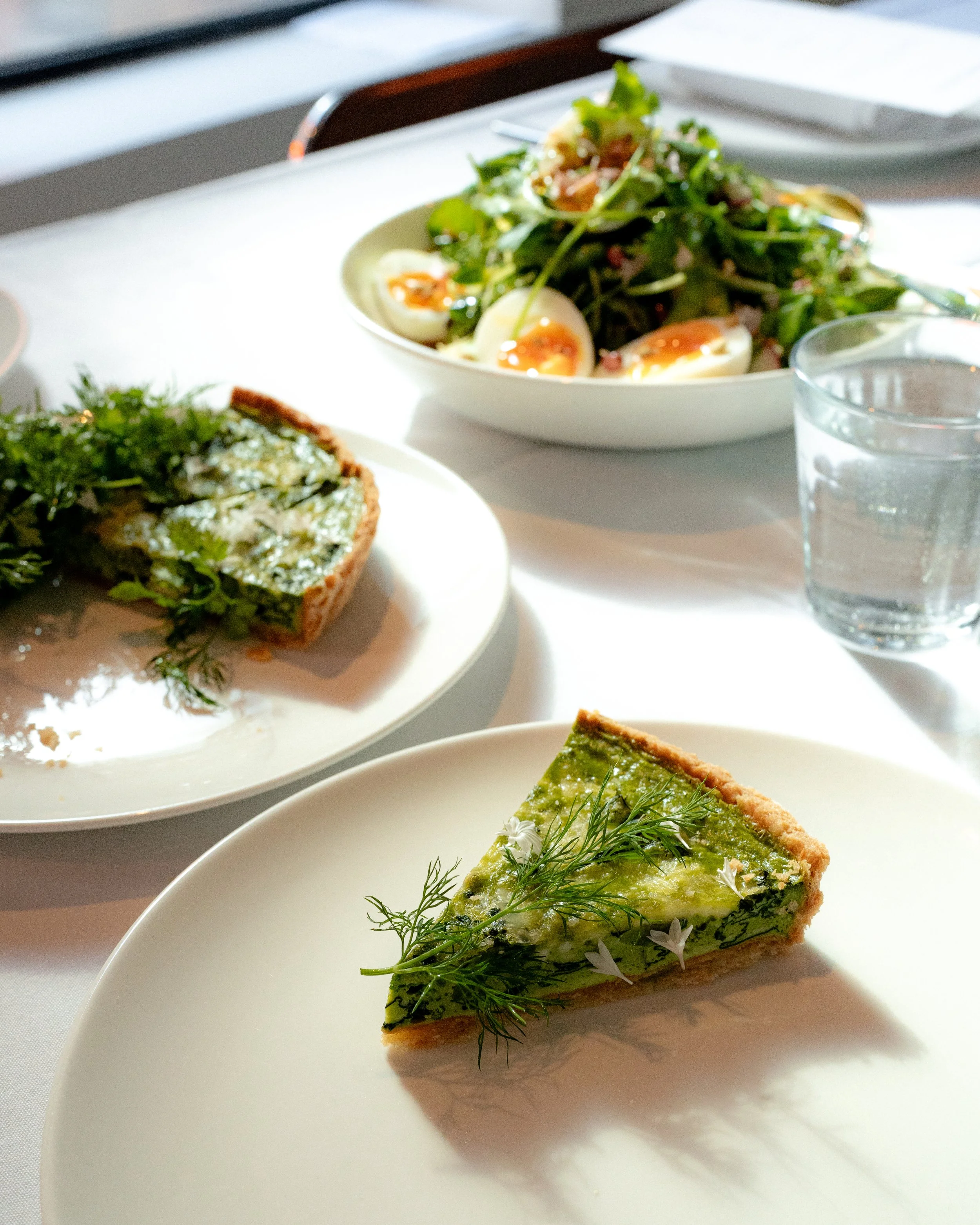 A slice of green vegetable quiche garnished with fresh dill and small white flowers on a white plate, with other dishes and a glass of water on a white table.