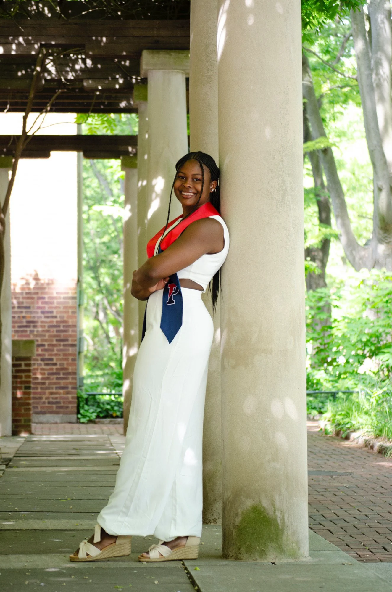A woman in a white dress with a red and white top, wearing beige platform wedge sandals, is standing outdoors under a pergola with green foliage in the background, smiling and leaning against a column.