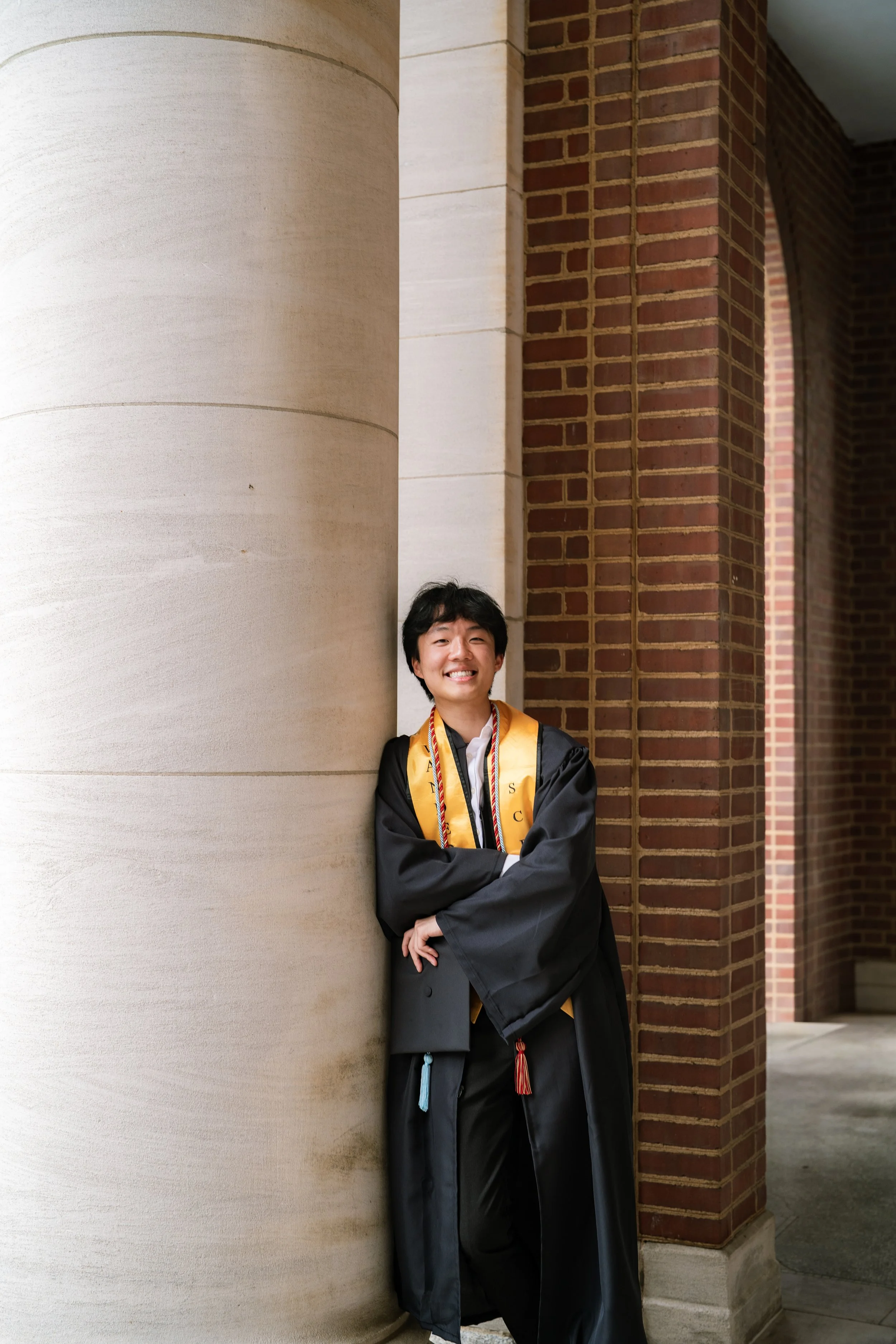 A young man in graduation attire, including a black cap and gown with gold and red cords, standing and smiling with arms crossed, leaning against a large beige column with brick archways in the background.
