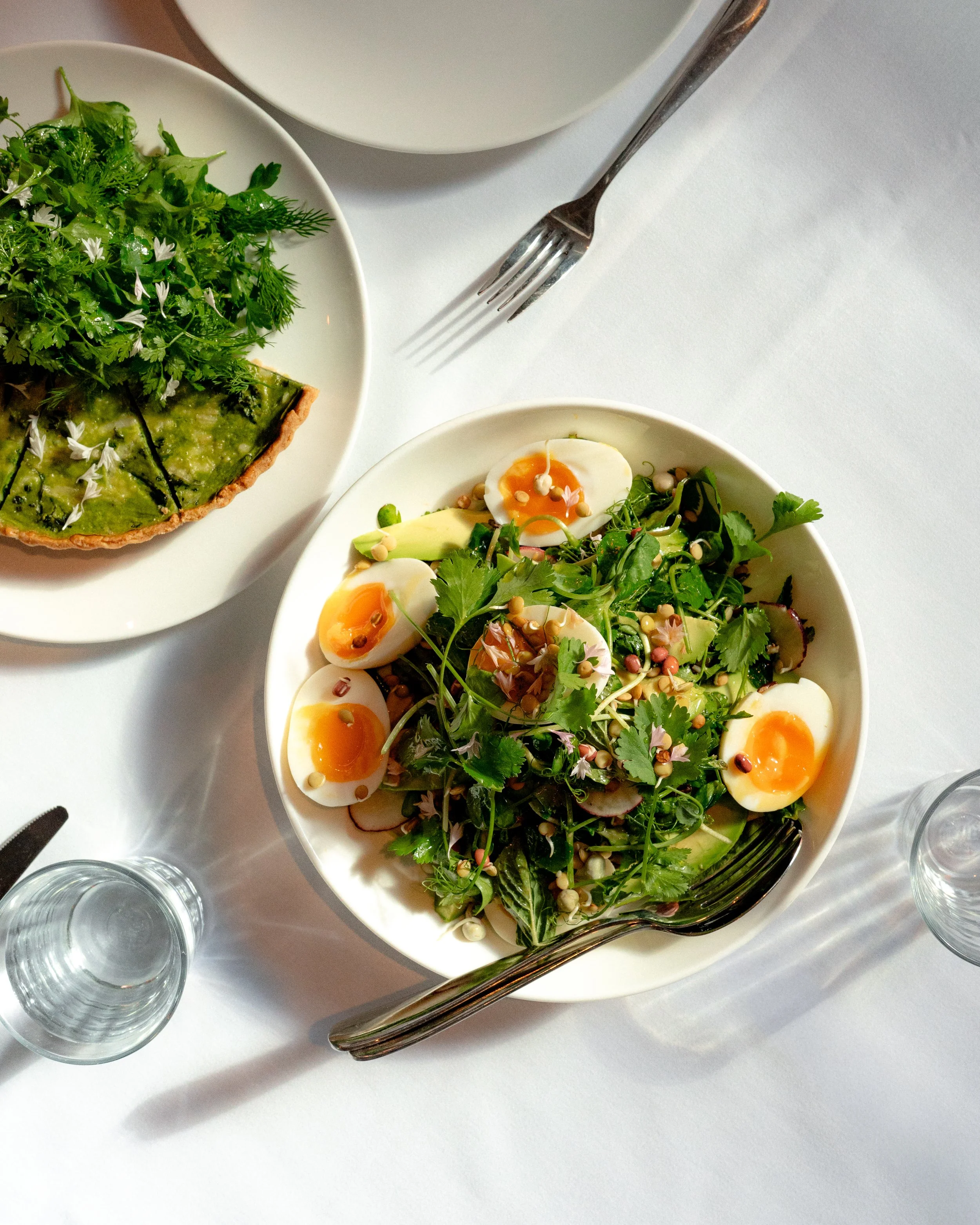 Overhead view of a table with a white tablecloth, featuring a bowl of salad with boiled eggs, greens, and edible flowers, a pie garnished with herbs and flowers, and two empty glasses of water.