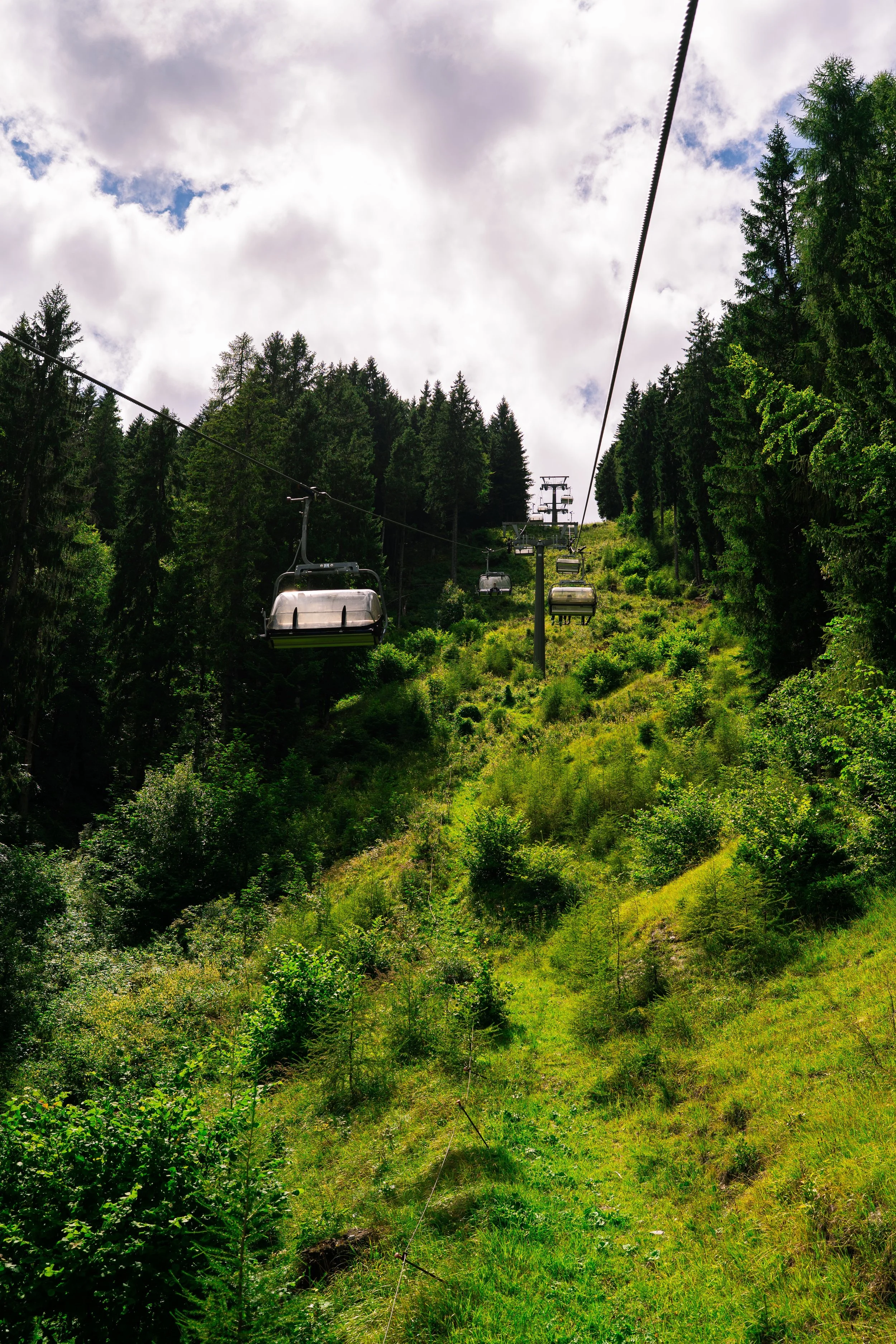A ski lift ascending through a forested mountain slope with green trees and cloudy sky.