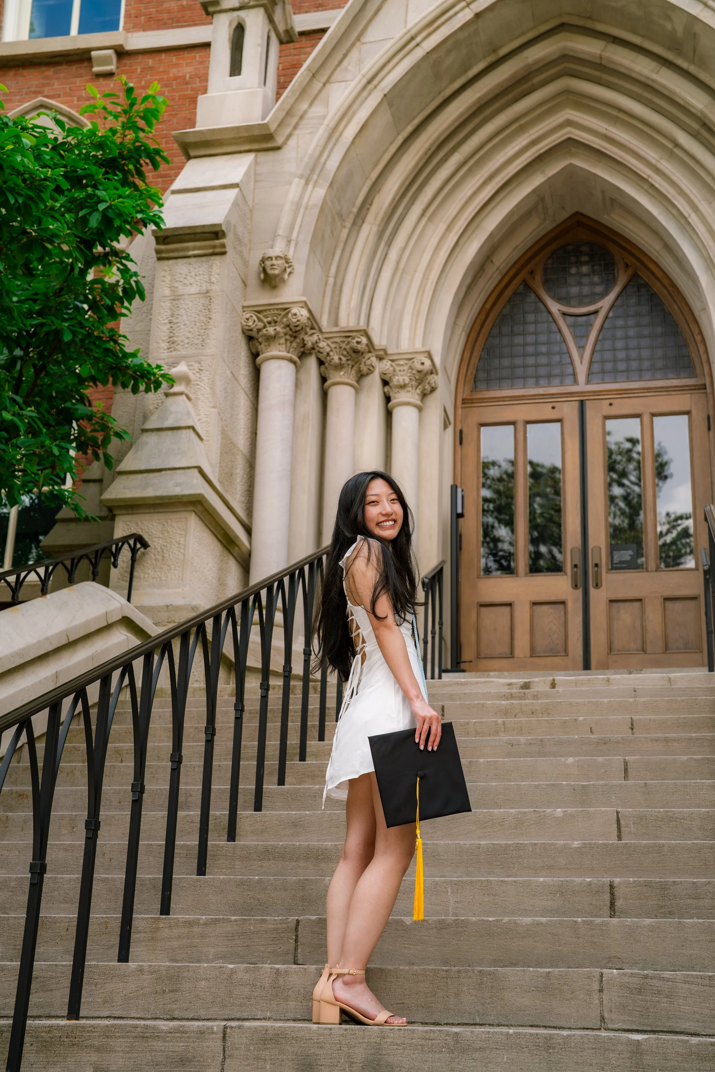 A young woman in a white dress, holding a diploma, smiling at the camera while standing on stone steps in front of a historic building with ornate architecture and gothic-style archway.