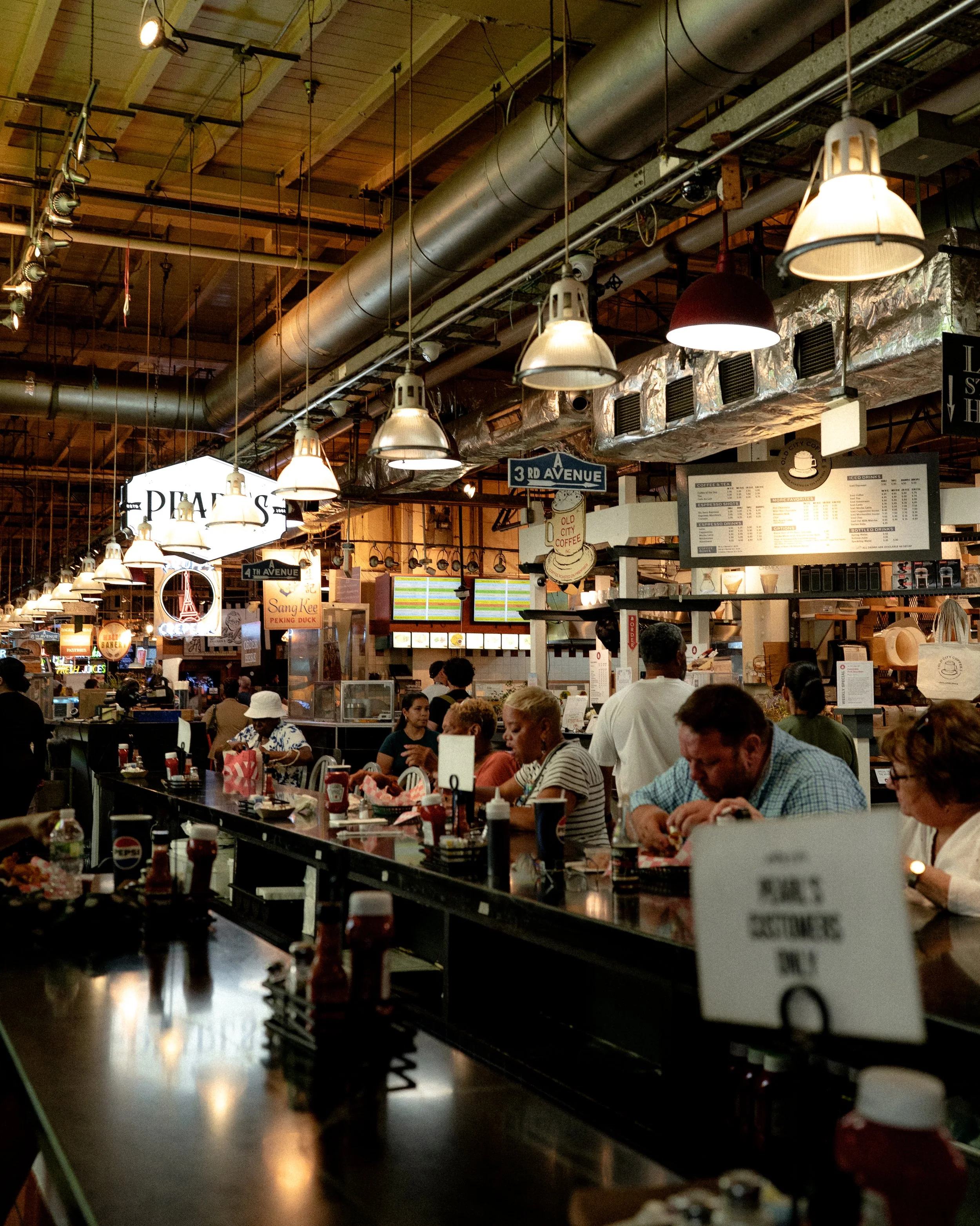 Inside Philadelphia's Reading Terminal Market, a busy food court, with people sitting and eating at tables and counters, illuminated by hanging lights, with a ceiling featuring exposed pipes and ductwork, and various signs and menus visible. 