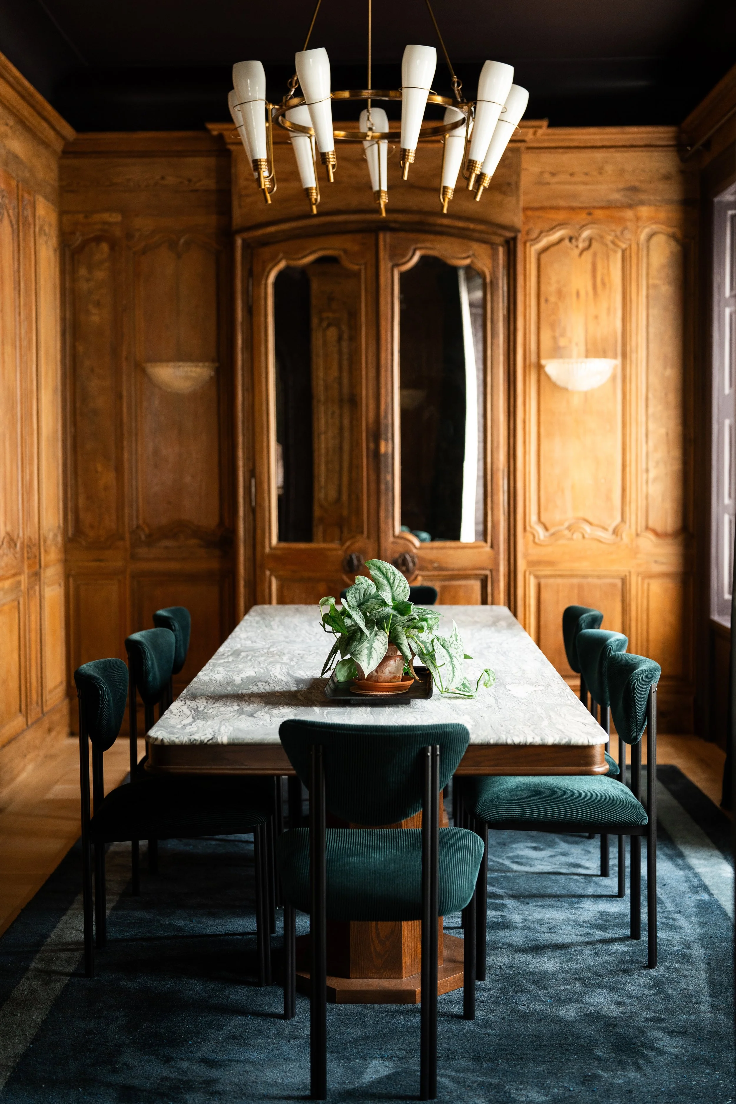 A dining room with a marble-top table and eight green velvet chairs. The table has a potted plant centerpiece. The room features wooden paneled walls, a large mirror, and a modern chandelier hanging above the table.