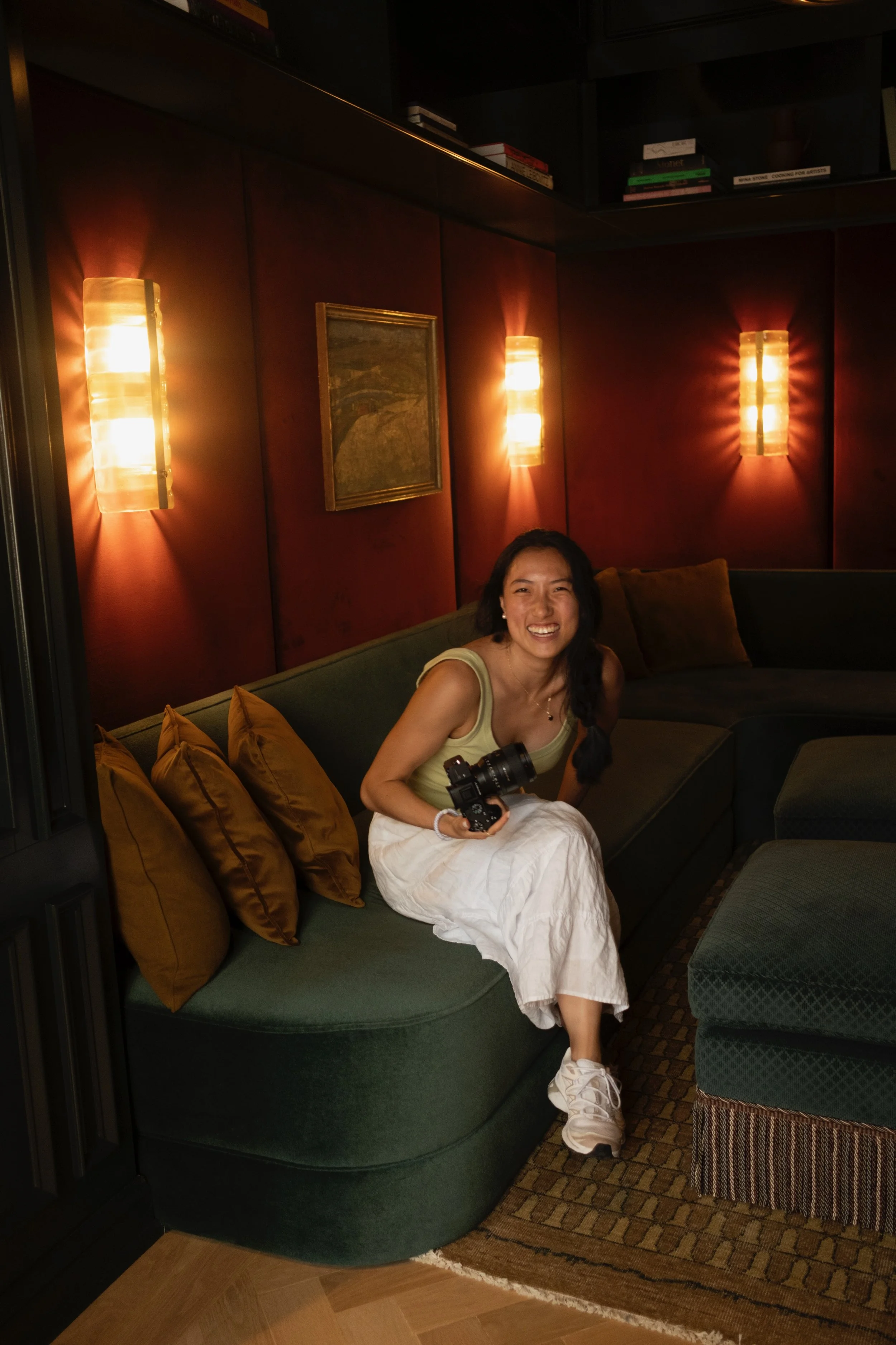A woman sitting on a green curved sofa with multiple brown pillows in a dimly lit room. She is smiling, holding a camera, wearing a yellow top and white skirt, with long dark hair. Behind her are red walls, framed artwork, and modern wall sconces casting warm light.