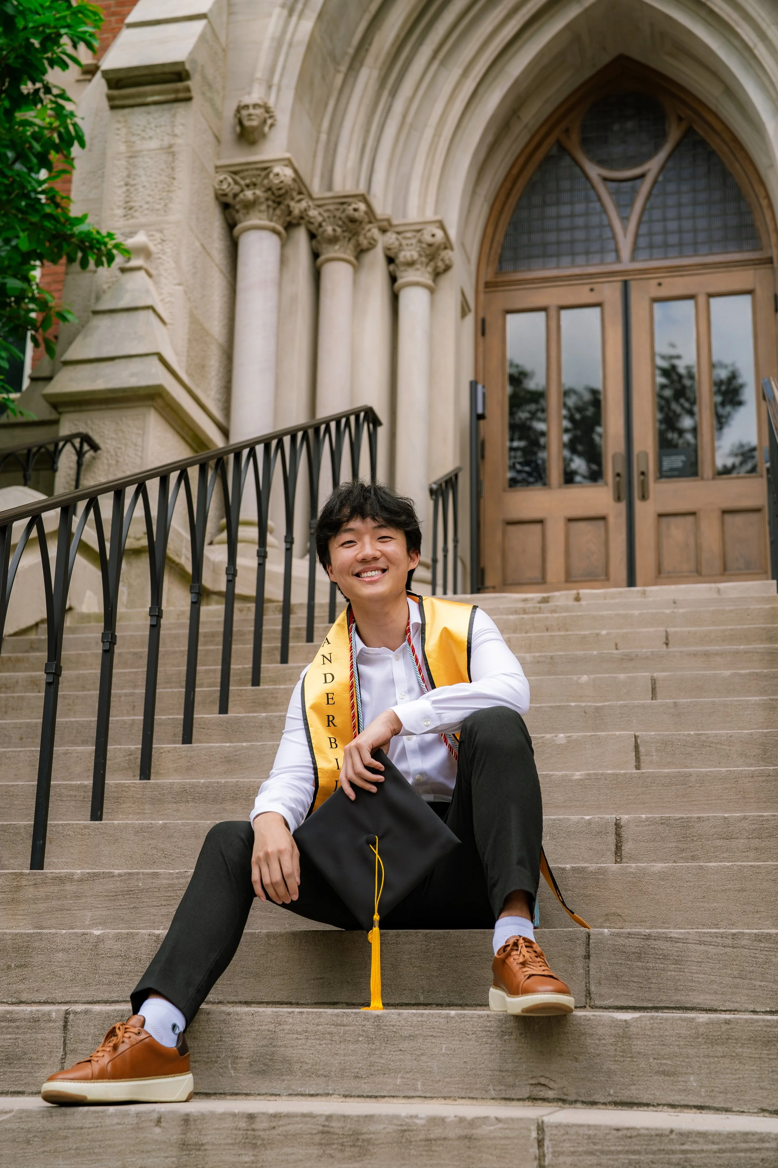 A young man sitting on stone steps in front of a large wooden door with windows, smiling, wearing a white shirt, black pants, brown sneakers, and a yellow graduation sash, holding a black cap with a yellow tassel.