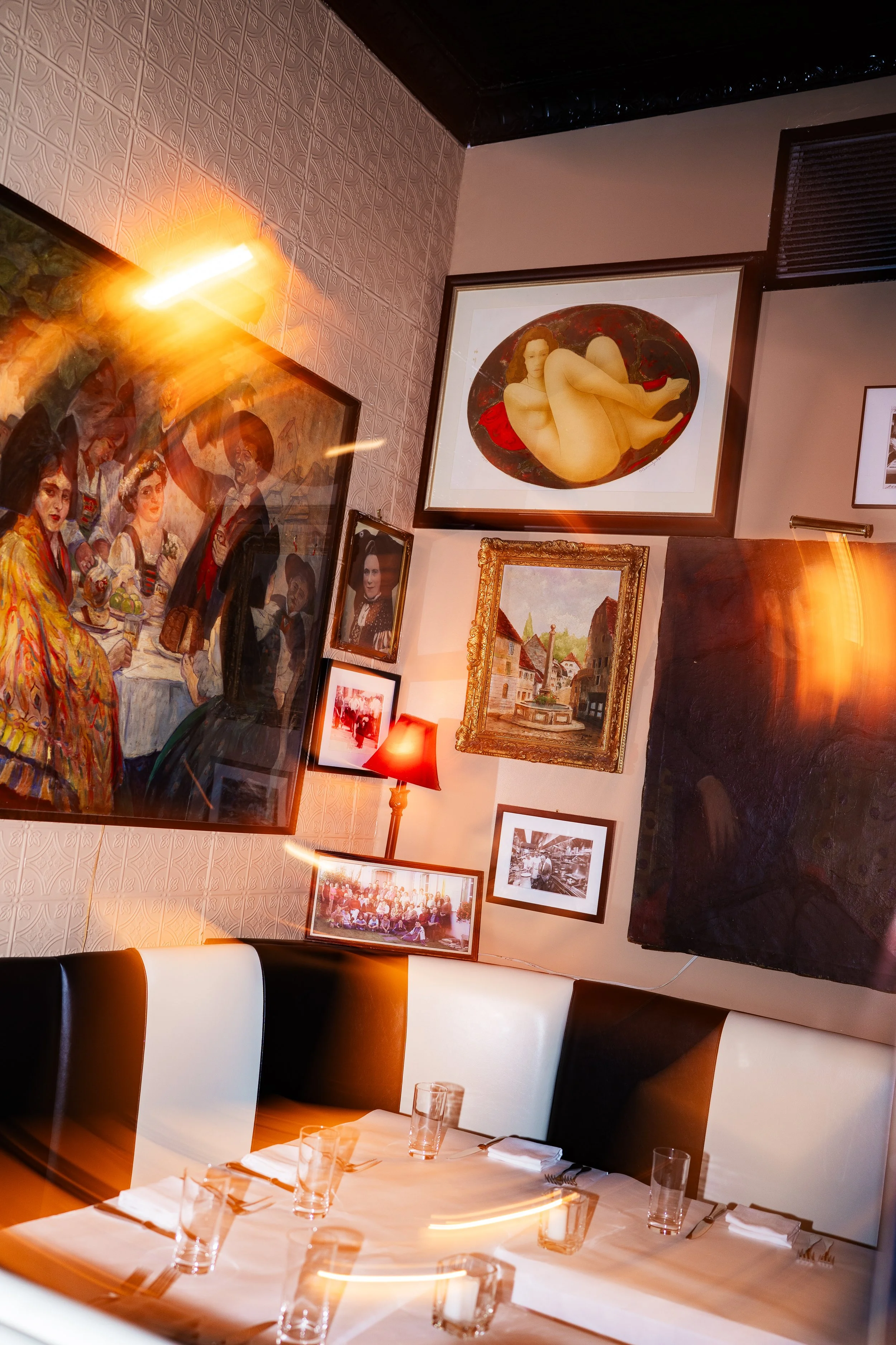 Interior of a restaurant with a dining table set with glasses and silverware, surrounded by black and white chairs, featuring a wall decorated with various framed paintings and photographs.