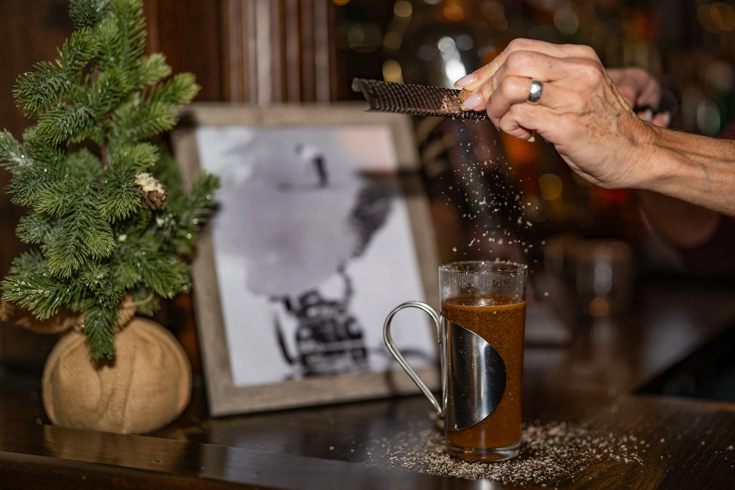 Person grating nutmeg over a glass of beer on a dark wooden table, with a small Christmas tree and decorative items in the background.