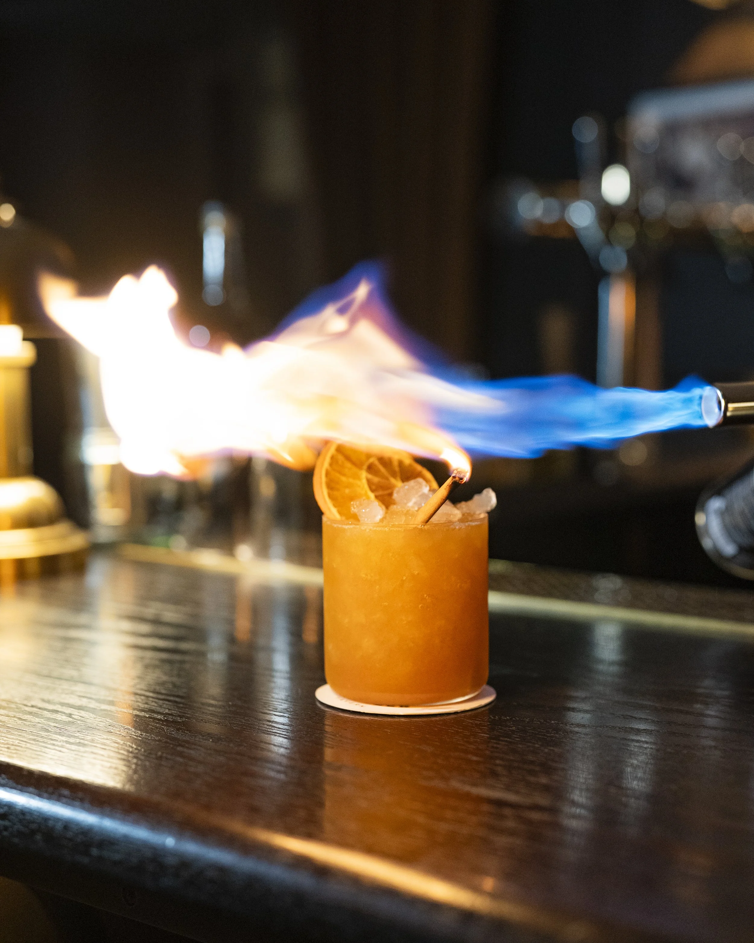 Colorful cocktail drink with orange slices and a flaming blue torch on top, served on a dark bar counter at night.