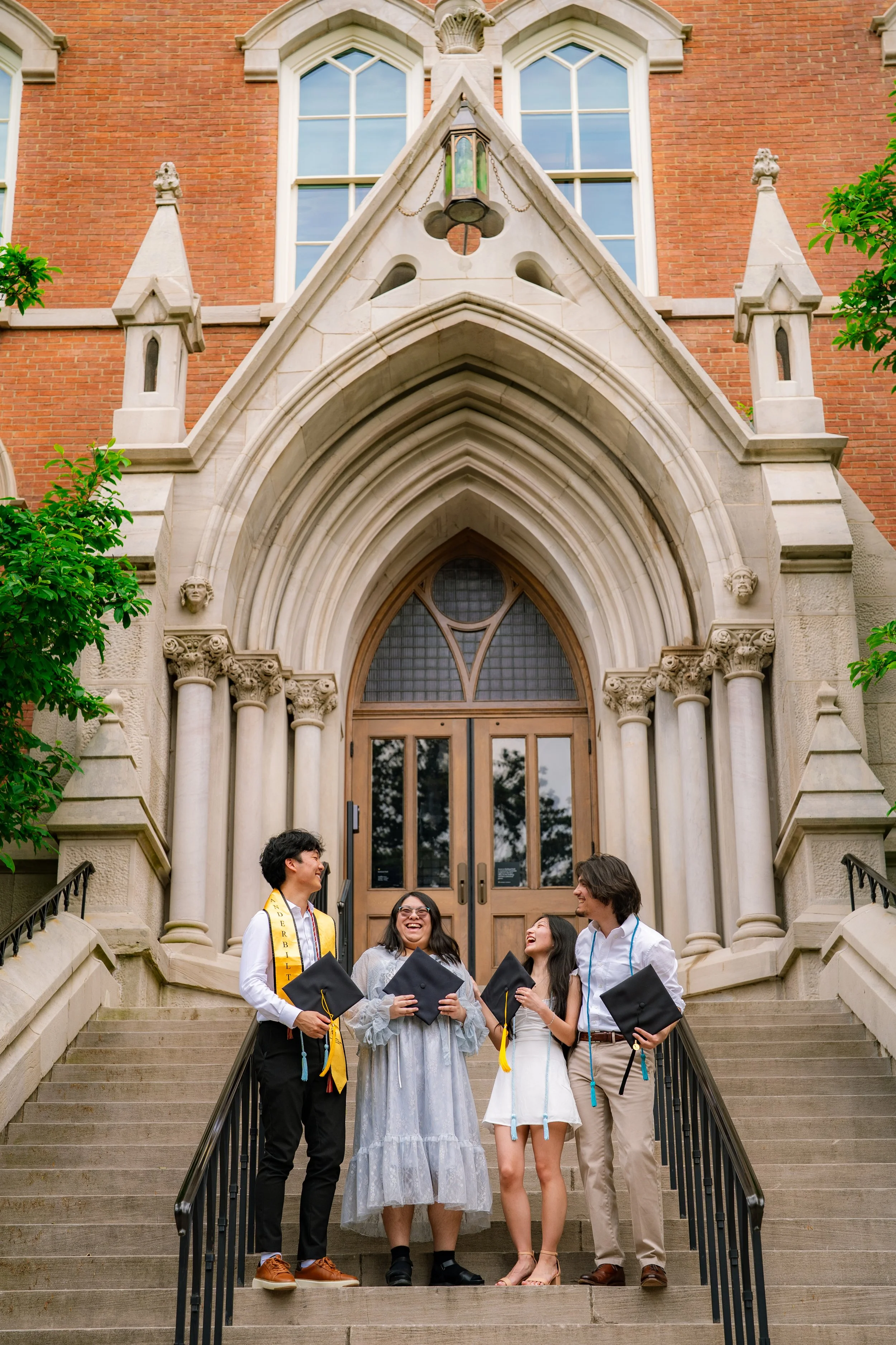 A group of four diverse graduation students standing on the steps of a cathedral-style building, wearing caps and gowns, smiling and talking.