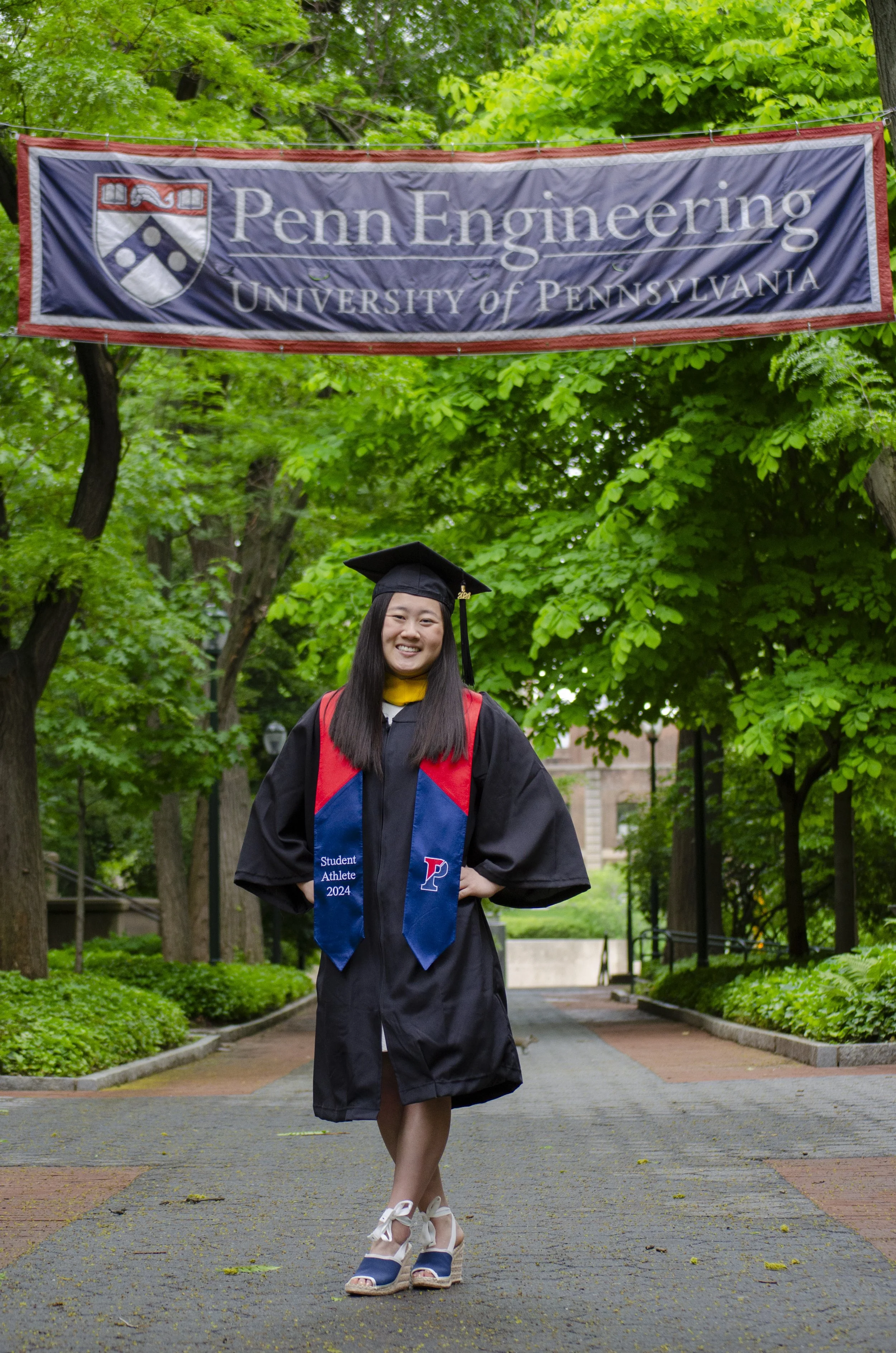 A young woman in a graduation cap and gown standing on a tree-lined pathway, smiling with her hands on her hips, with a Penn Engineering banner hanging above her.