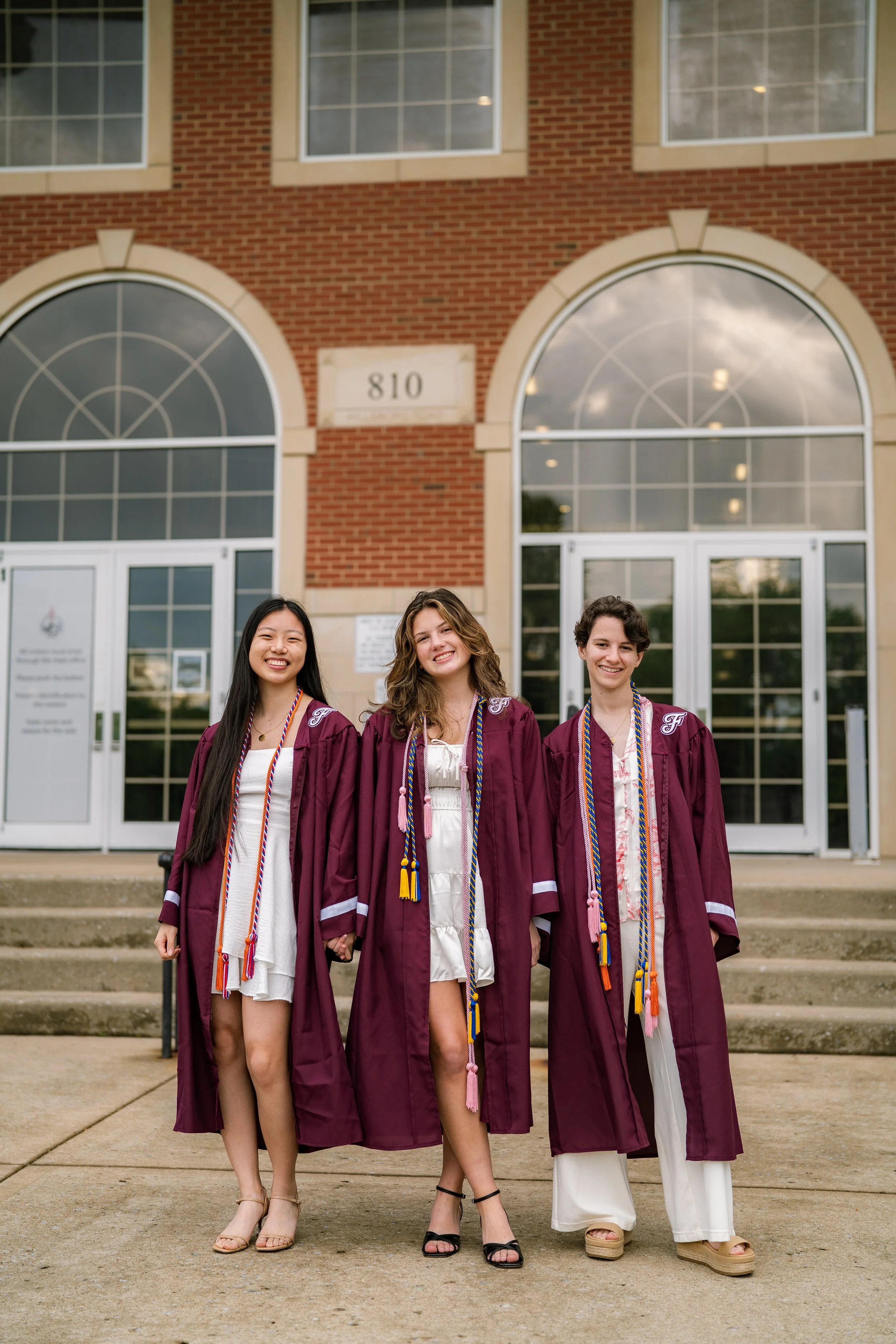 Three young women in maroon graduation gowns standing on steps in front of a brick building with large windows, smiling.