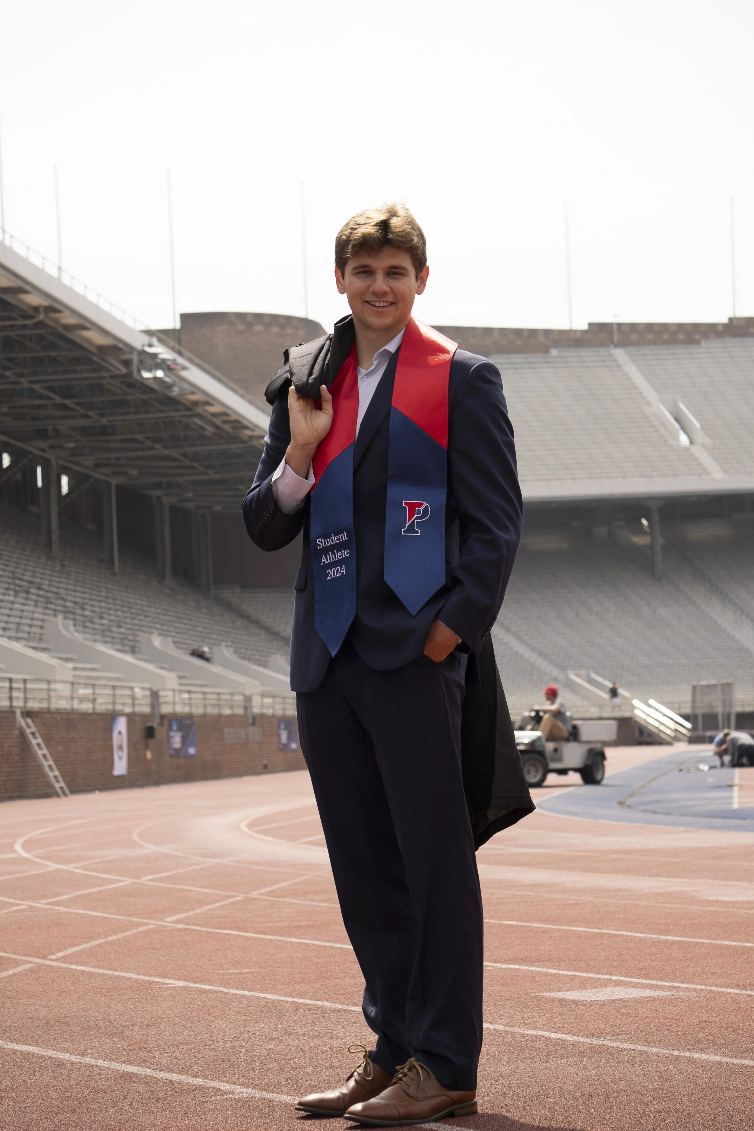 A young man in a suit standing on a running track, smiling, carrying a backpack, wearing a red, blue, and white sash that reads 'Student Athlete 2024' at a stadium.