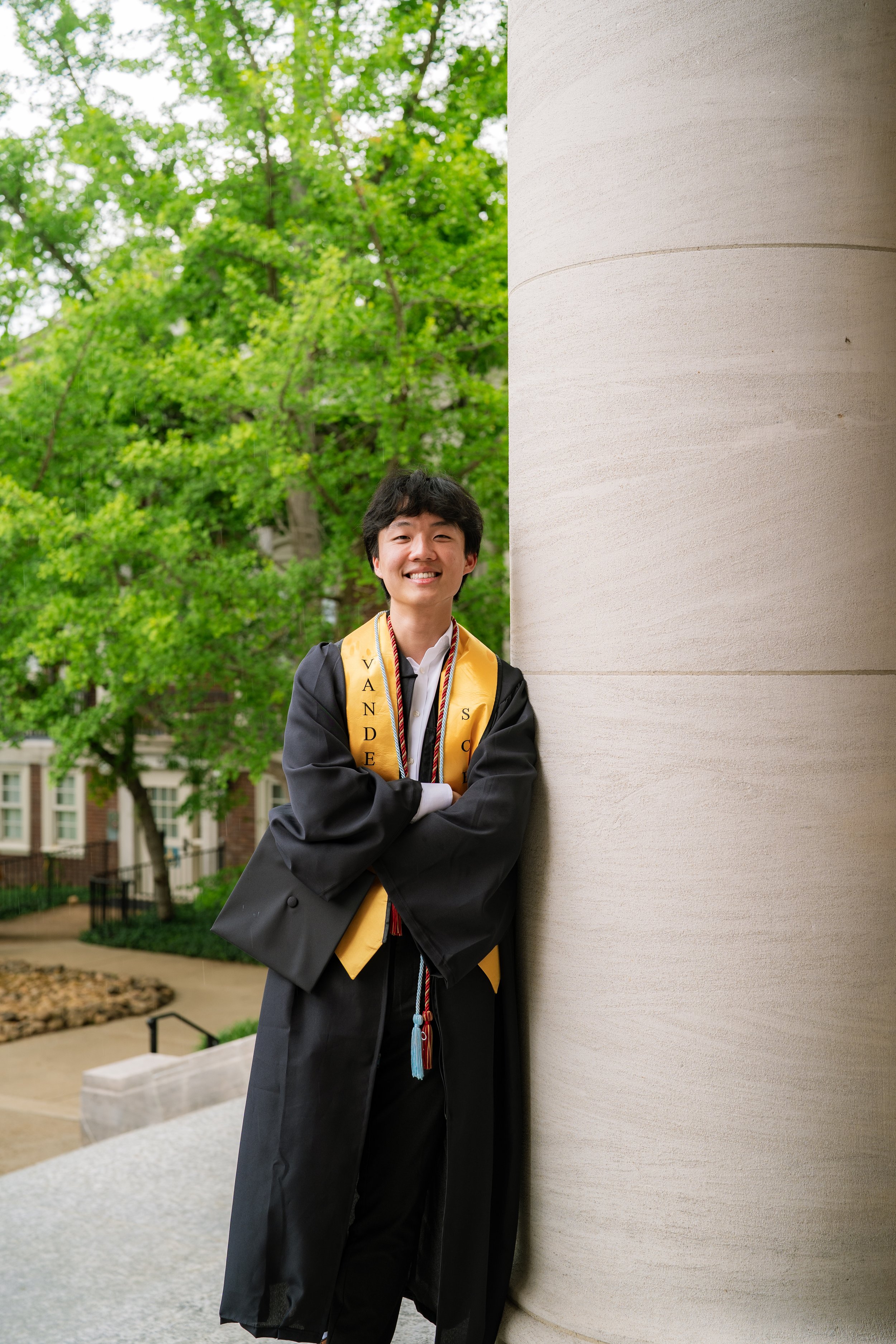 A young man wearing a graduation cap and gown, standing outdoors with arms crossed, smiling, next to a large stone column with green trees in the background.