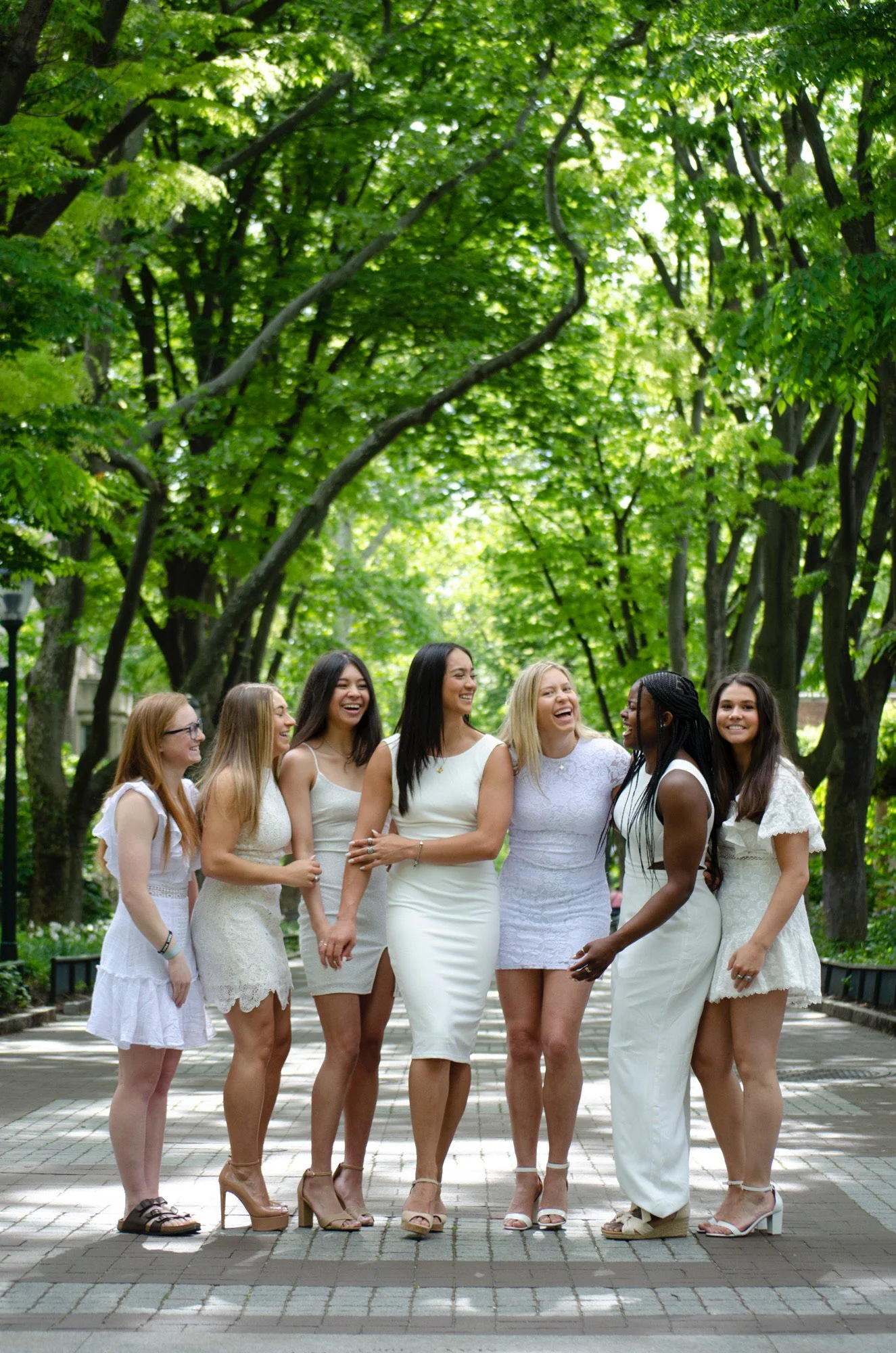 A group of diverse women in white dresses walking and smiling in a park with green trees overhead.