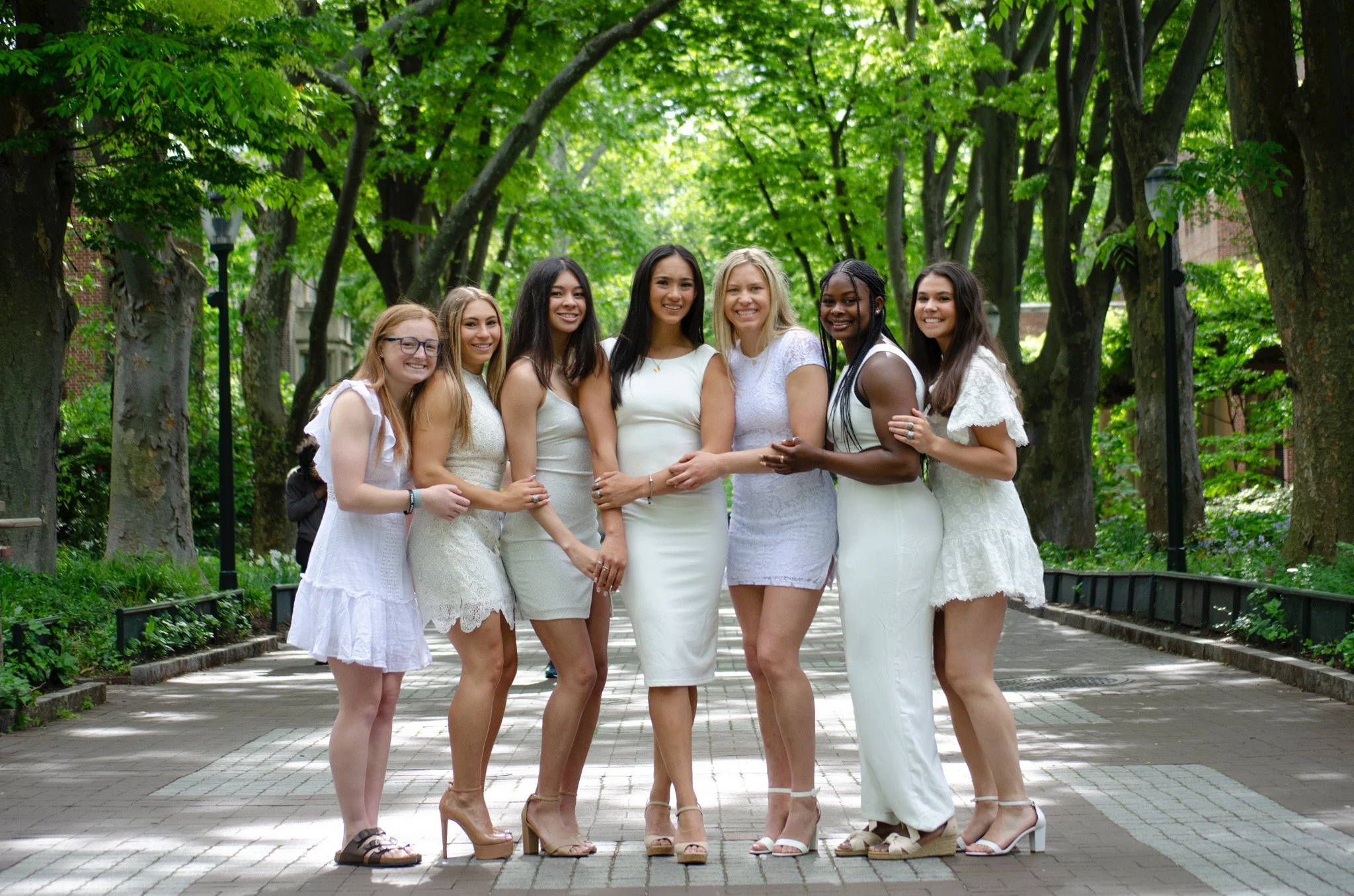 A group of nine diverse women standing arm in arm on a park path surrounded by green trees, smiling at the camera.