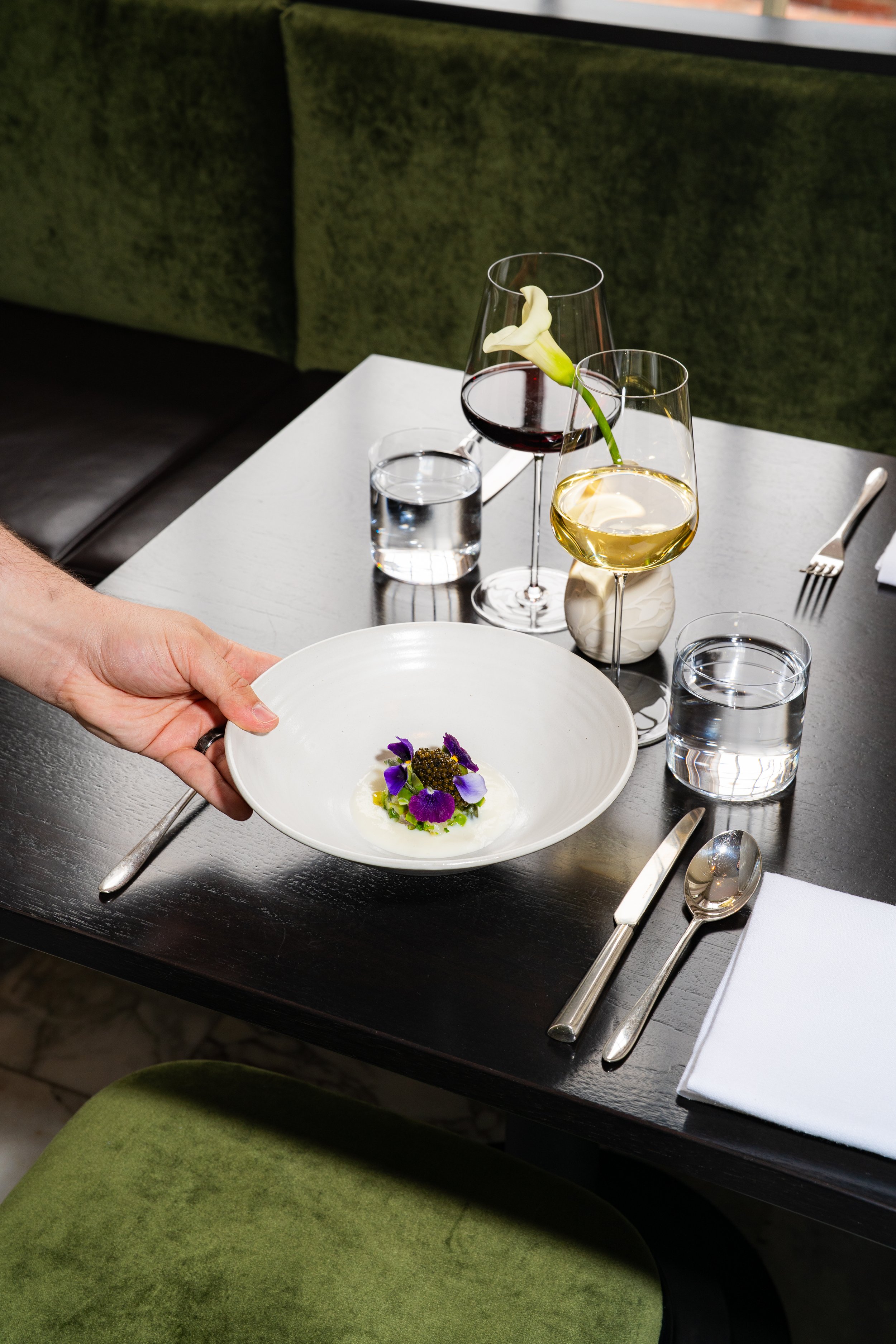 A person is holding a white plate with a gourmet dish garnished with purple edible flowers. In the background, there are two glasses of wine, one red and one white, and glasses of water on a dining table set with silverware, a fork, spoon, and a napk