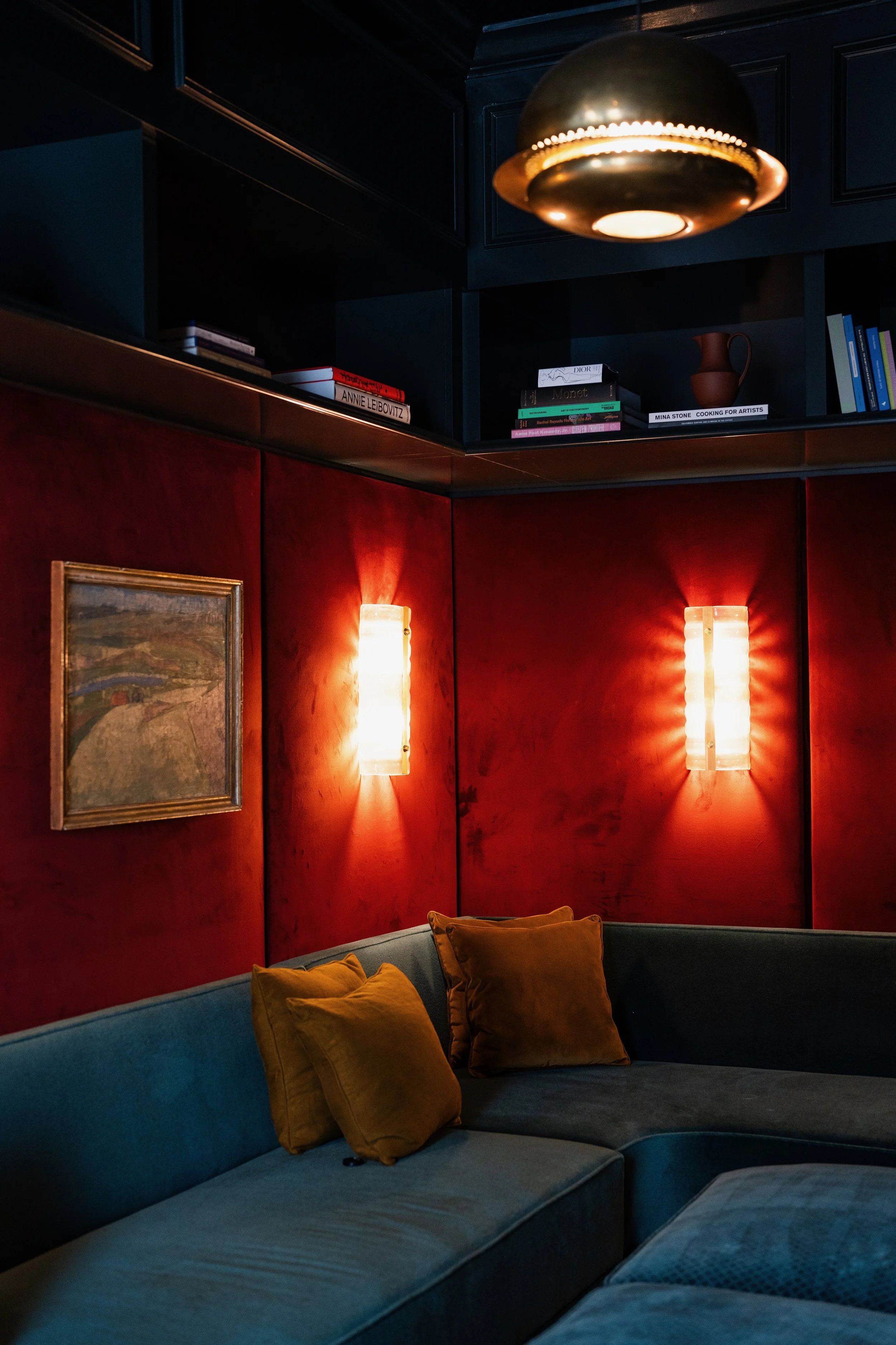 A corner of a room with a blue and red sectional sofa with yellow cushions, red walls, wall-mounted lights, a framed artwork on the red wall, and black shelving above with books, a small brown pitcher, and a gold and black ceiling light fixture.
