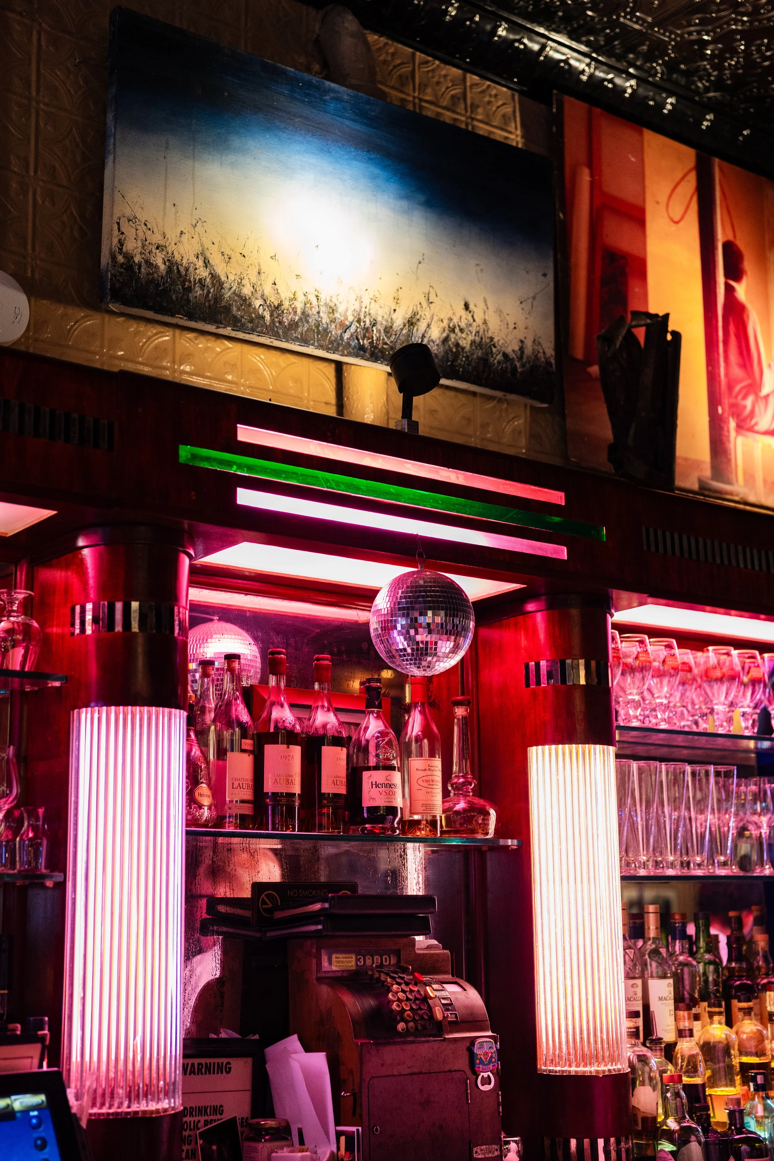 A bar with bottles of liquor, illuminated by pink and purple lighting, with two vertical light fixtures, a disco ball, and a portrait painting on the wall.