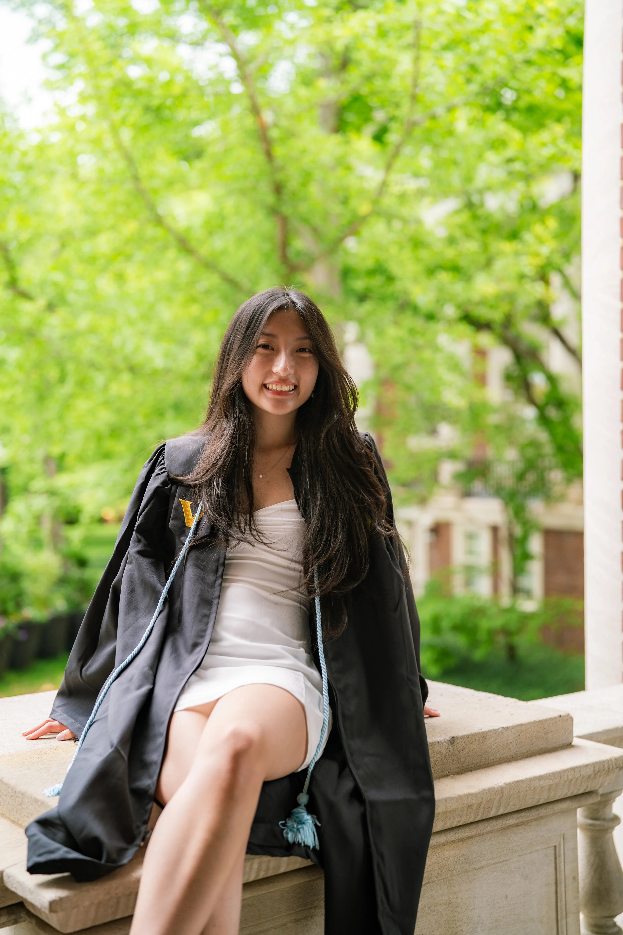 A young woman in a graduation cap and gown sitting on a stone ledge outdoors, smiling at the camera, with green trees in the background.