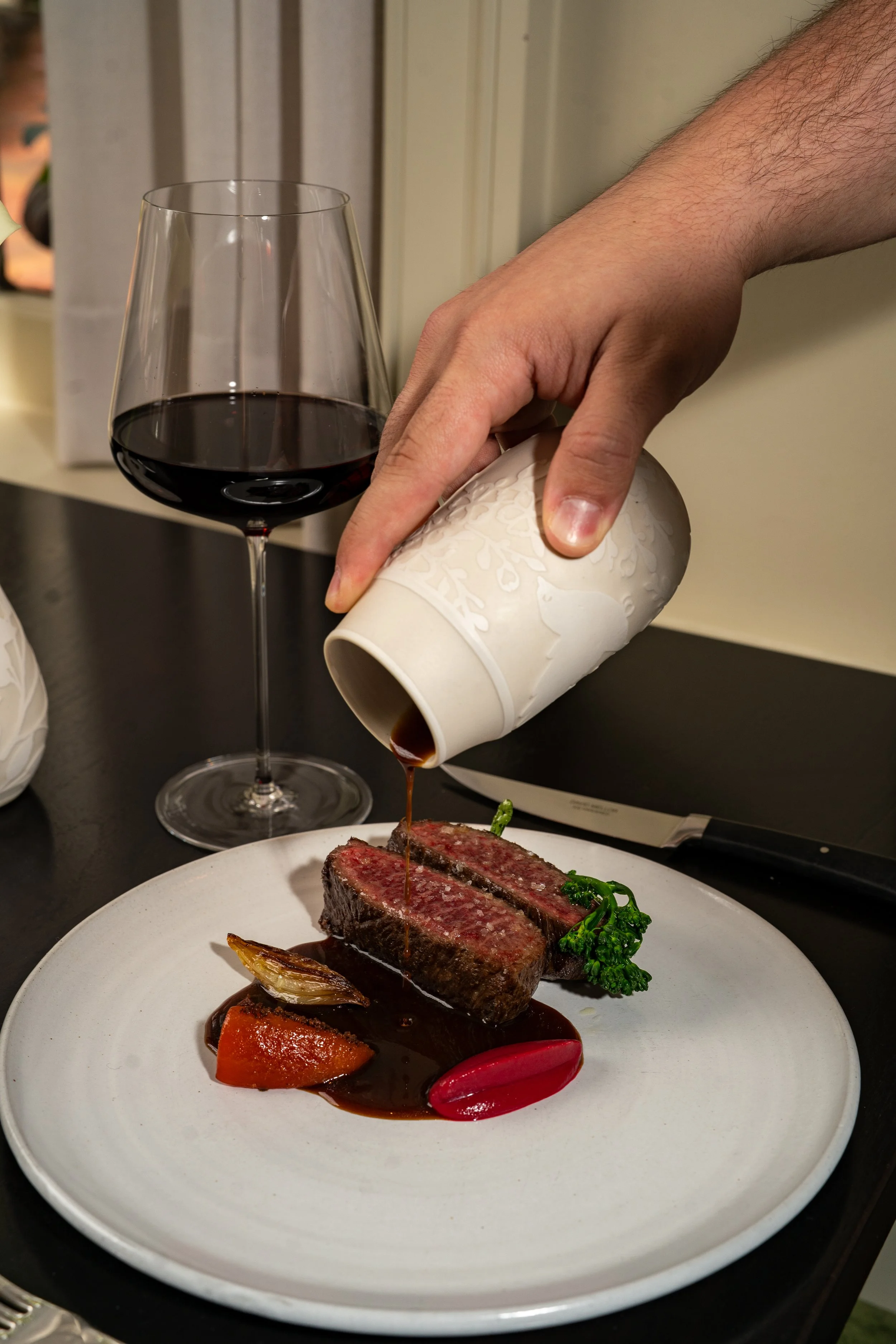 A hand pouring sauce over a cooked steak with vegetables on a white plate, with a glass of red wine nearby.