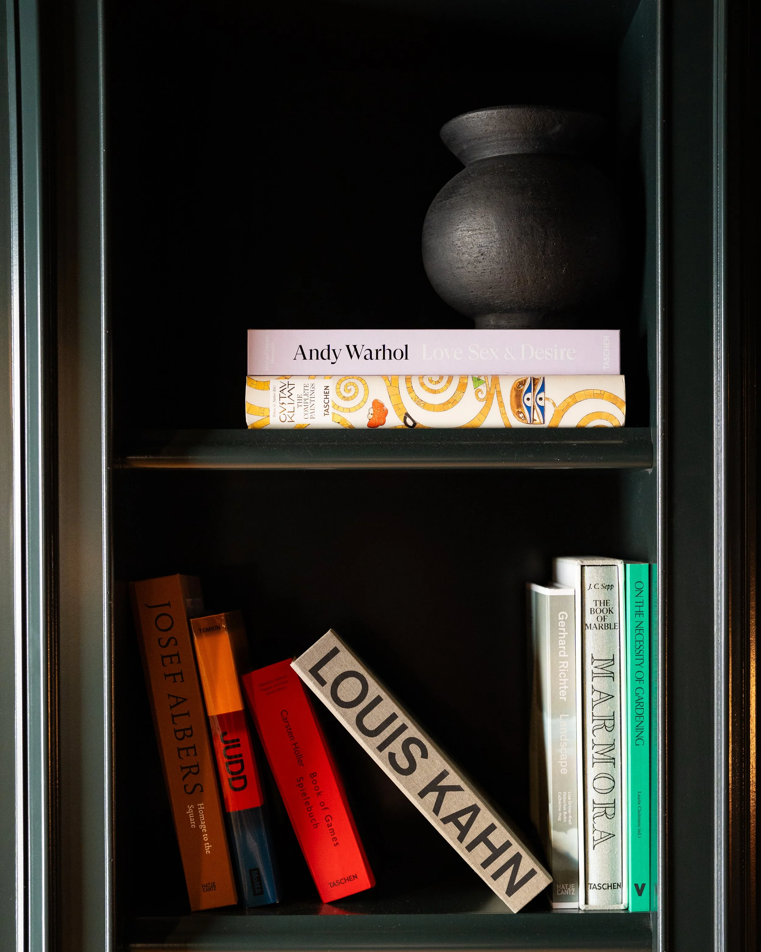 A black bookshelf with books, including a pink book titled "Love Sex & Desire" by Andy Warhol, an art book with a colorful cover, and various other books with titles on topics like art, architecture, and gardening.