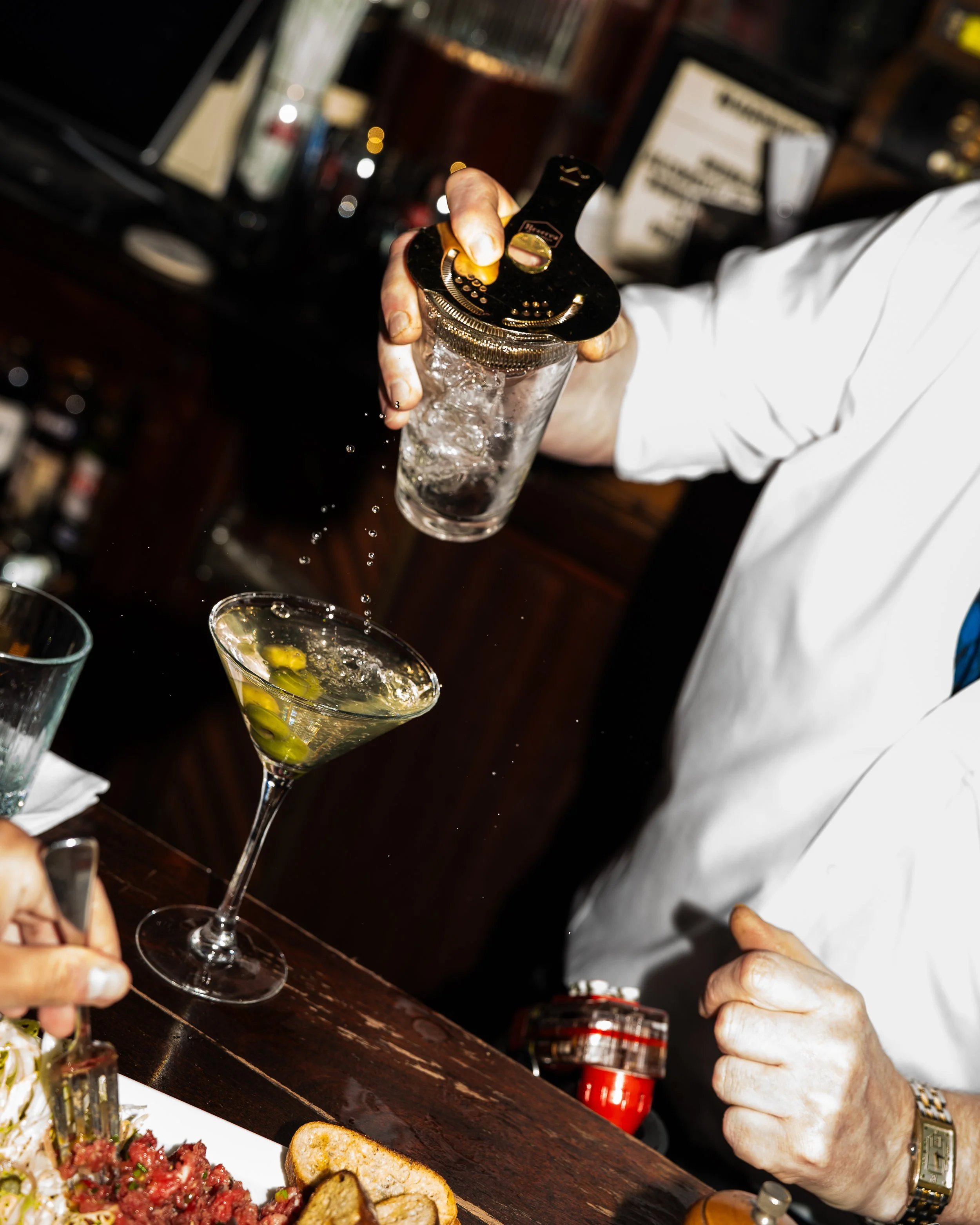 Person pouring a clear liquid into a glass with ice and olives, with food and drinks on the table nearby.