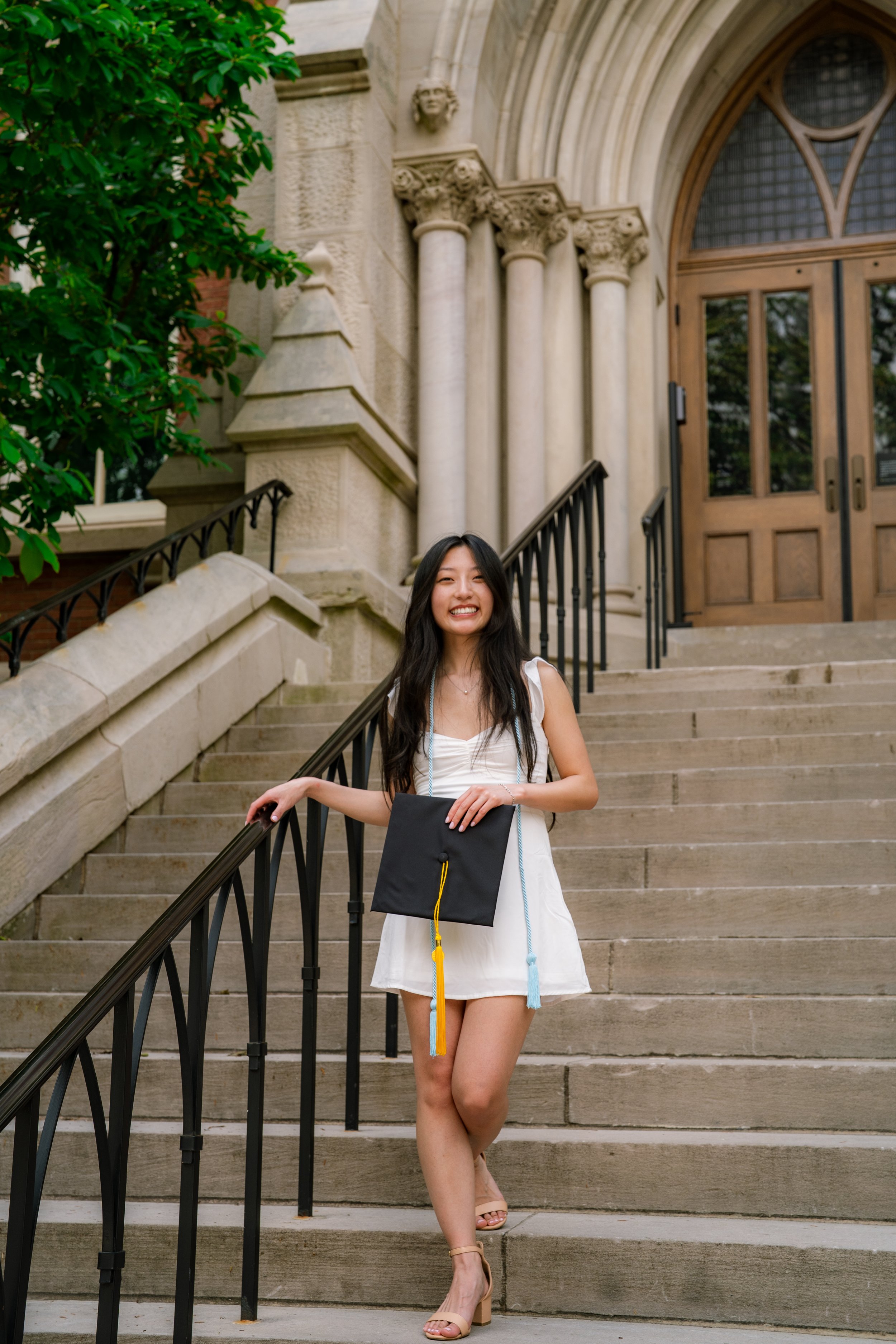 A young woman in a white dress standing on stairs in front of an ornate stone building, holding a black graduation cap with tassels, smiling at the camera.