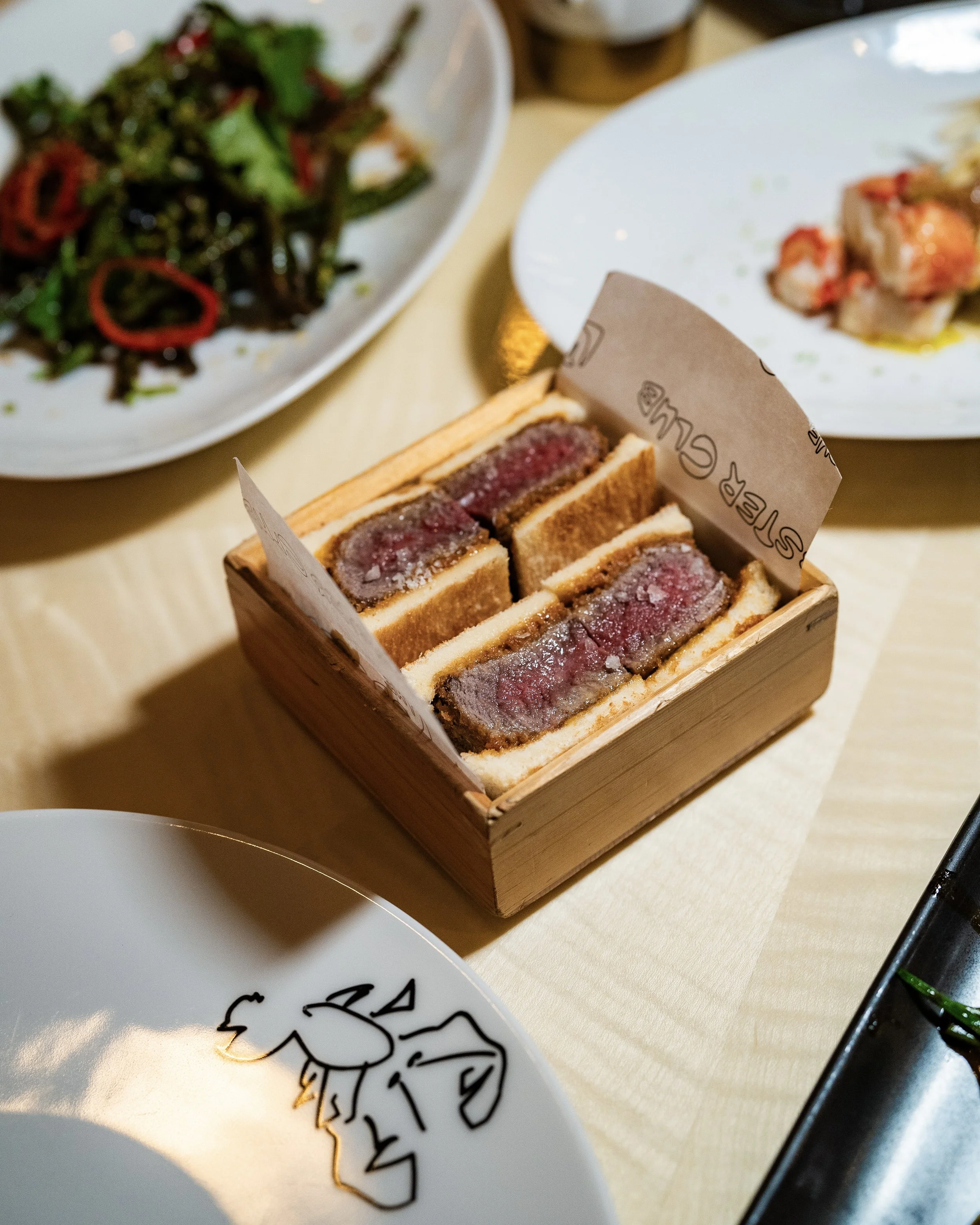 A wooden box of sliced beef Wellington on a table, with side dishes of salad and possibly seafood. The table has plates and a glass bottle.