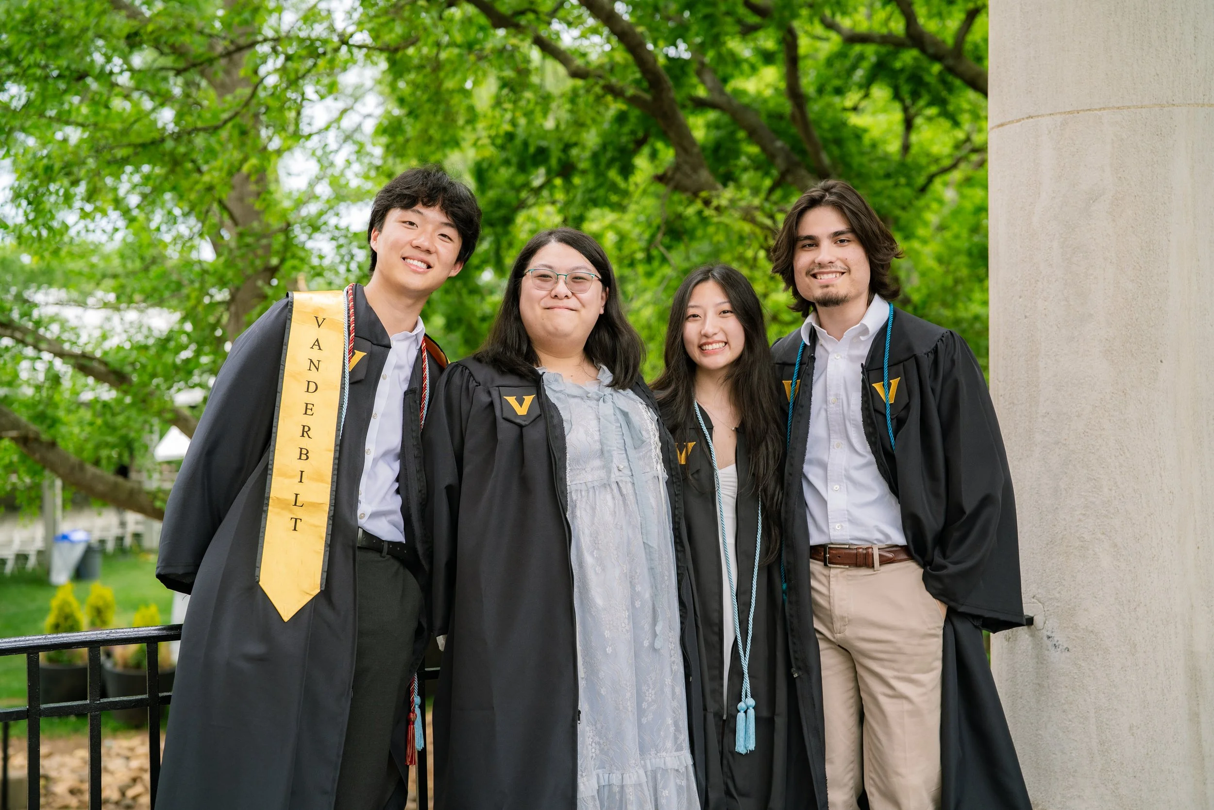 Four diverse young adults in graduation caps and gowns smiling outdoors, celebrating graduation, with green trees in the background.