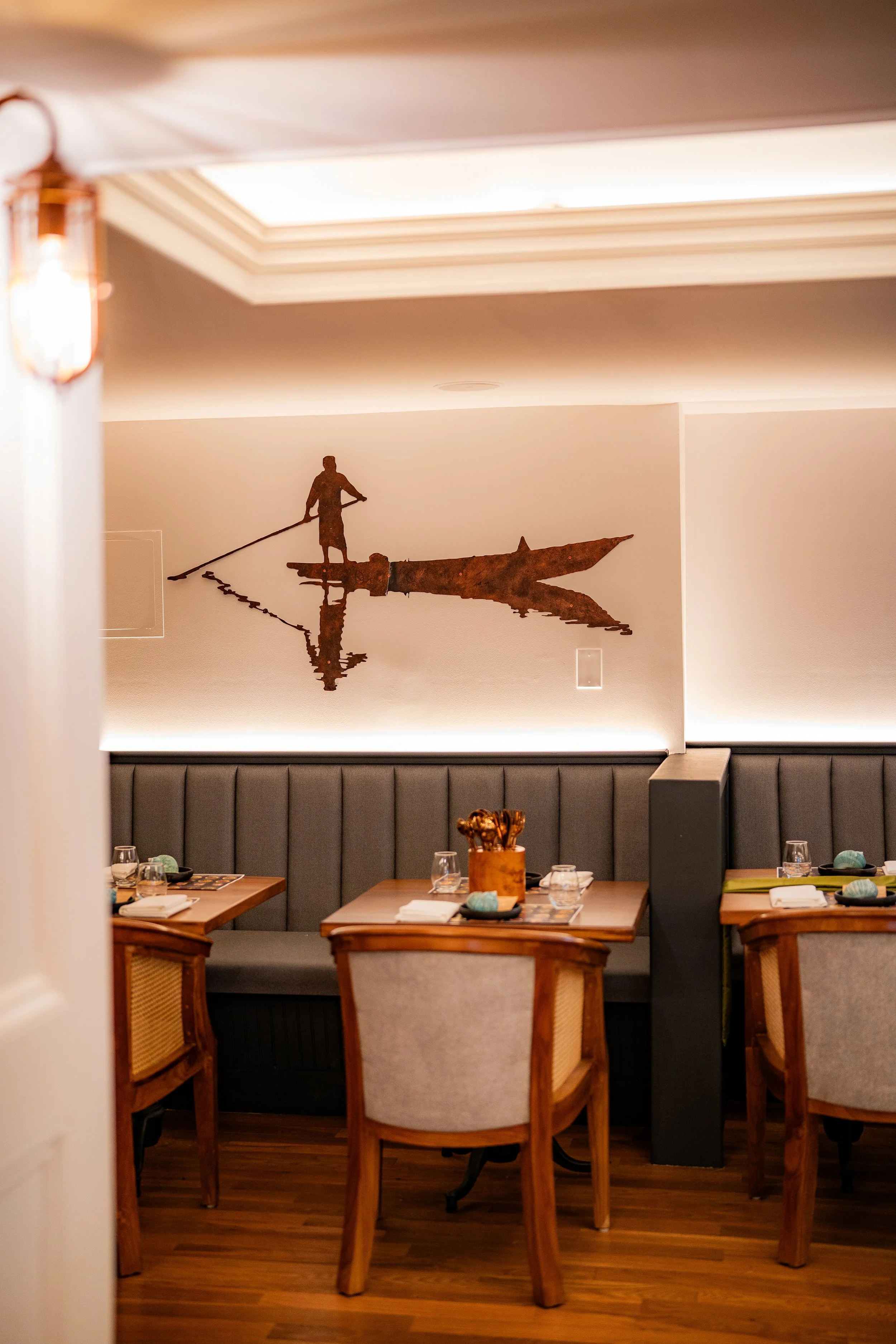 A restaurant dining area with wooden tables and chairs, and a gray upholstered banquette. There is a wall decoration of a silhouette of a person standing in a boat, holding a pole, reflected on the water. The room has warm lighting and wooden floorin