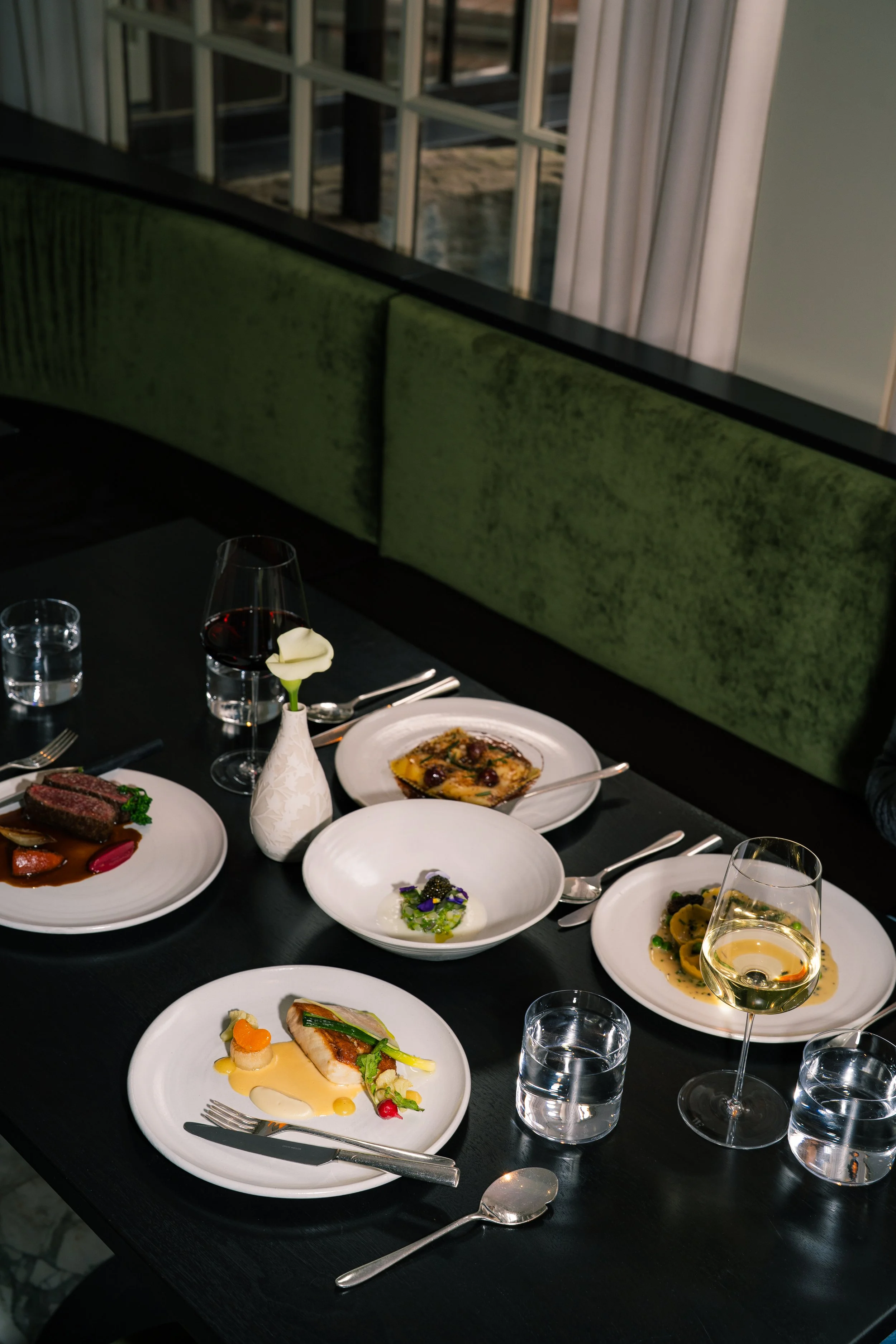 A table set with various gourmet dishes in a restaurant, including plates of meat, fish, and vegetables, along with a glass of white wine, a glass of water, and utensils.