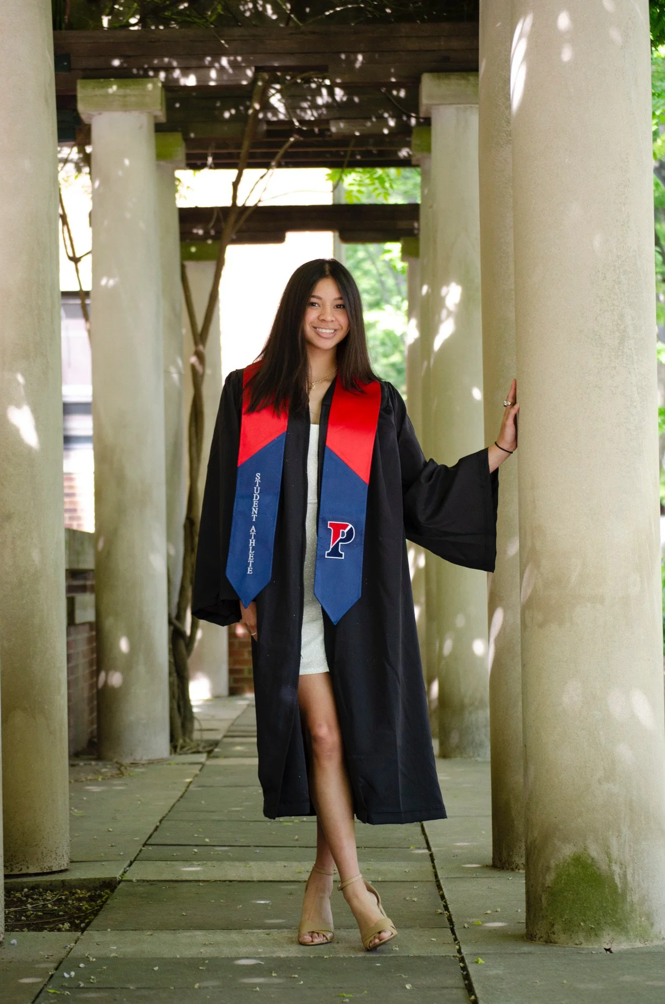 Young woman in graduation gown and hood standing among columns outdoors, smiling, with one hand on a column, and wearing heels.