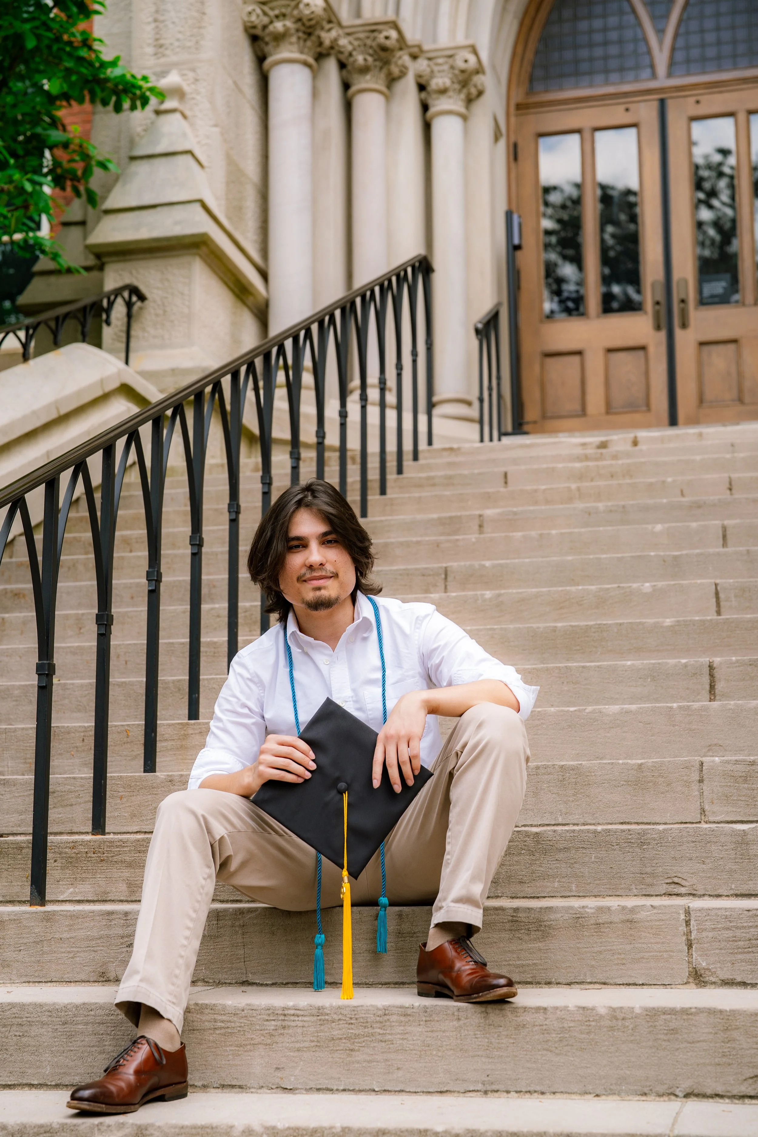 A young man with long hair, facial hair, and wearing a white shirt, beige pants, and brown shoes, sitting on outdoor steps in front of a building with large wooden doors and columns, holding a black graduation cap with a yellow tassel.