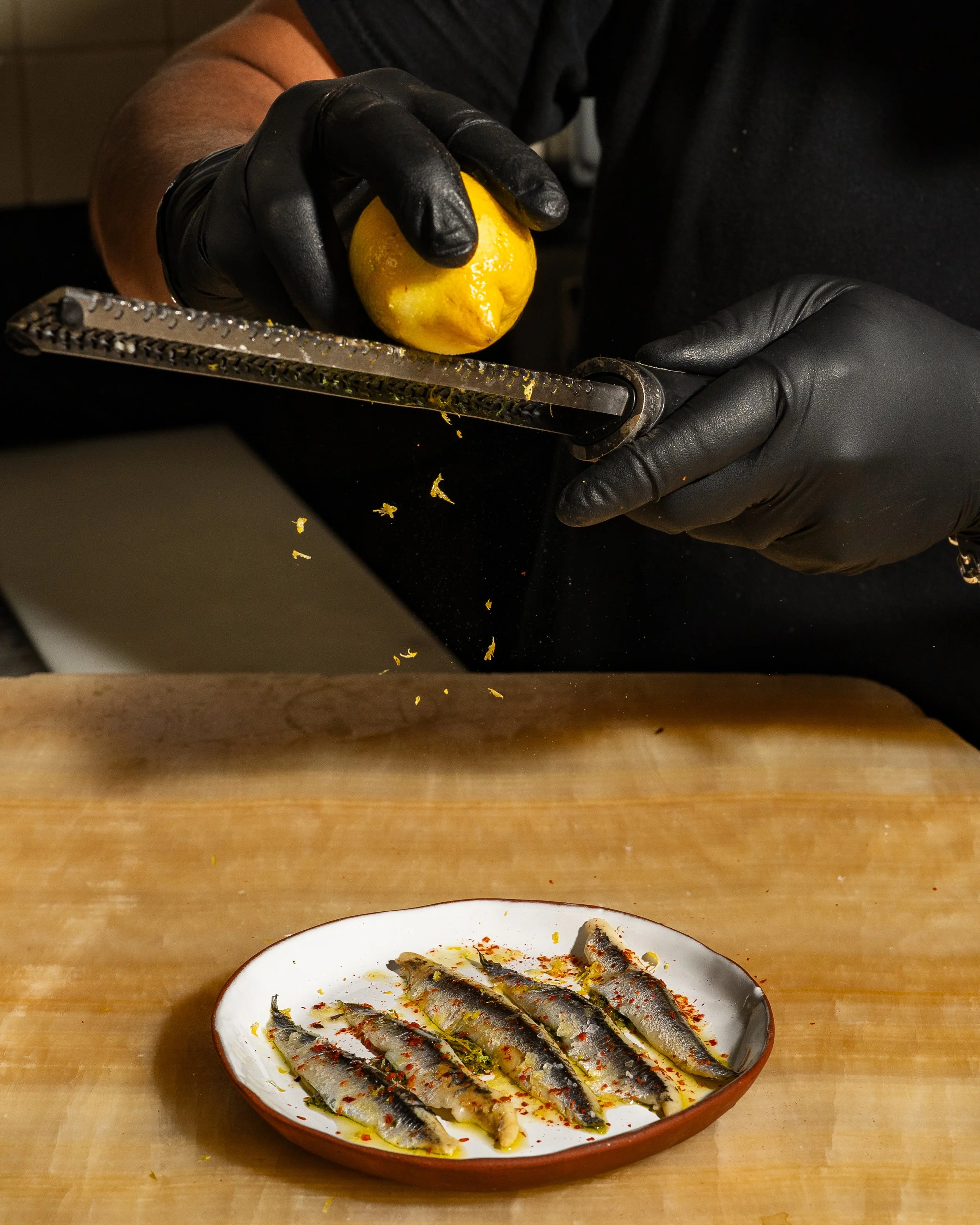 A person wearing black gloves is grating a lemon over a dish of grilled sardines, which are served on a white plate with red spices.