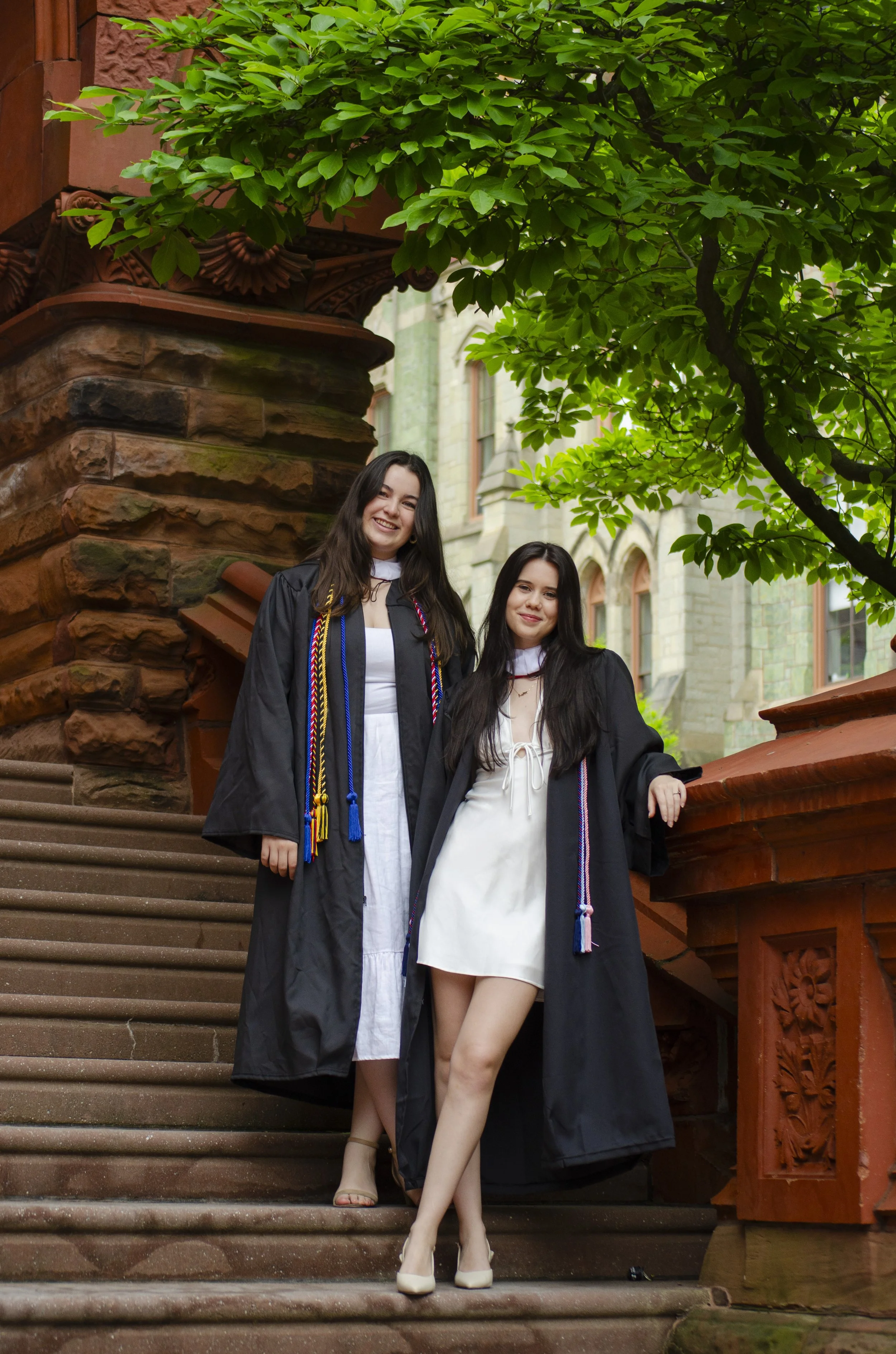 Two young women in graduation robes standing on the stairs of a historic building with intricate stonework, green trees, and windows in the background.