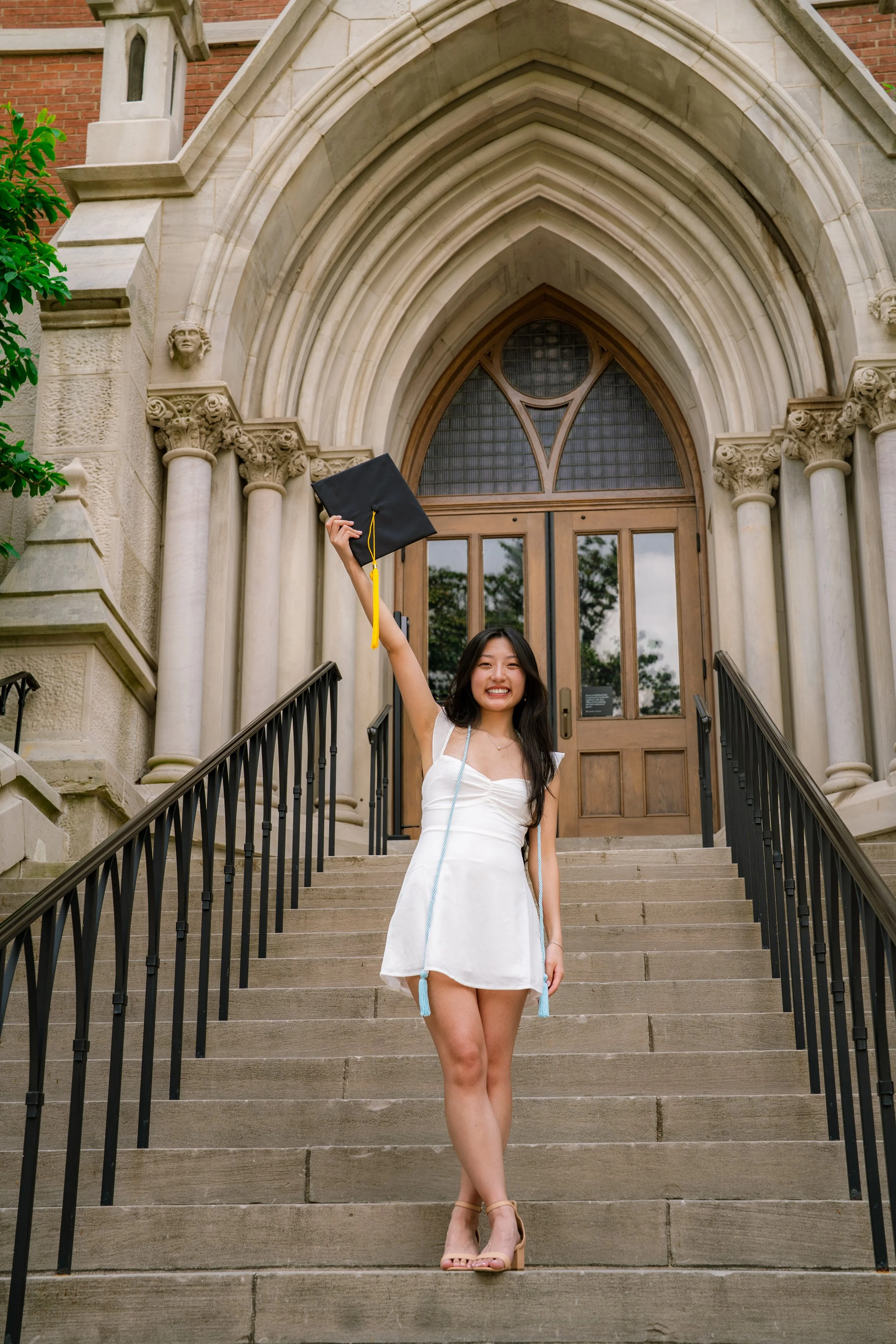 A young woman in a white dress is standing on outdoor stairs in front of a church with Gothic architecture, holding her graduation cap high in the air and smiling.