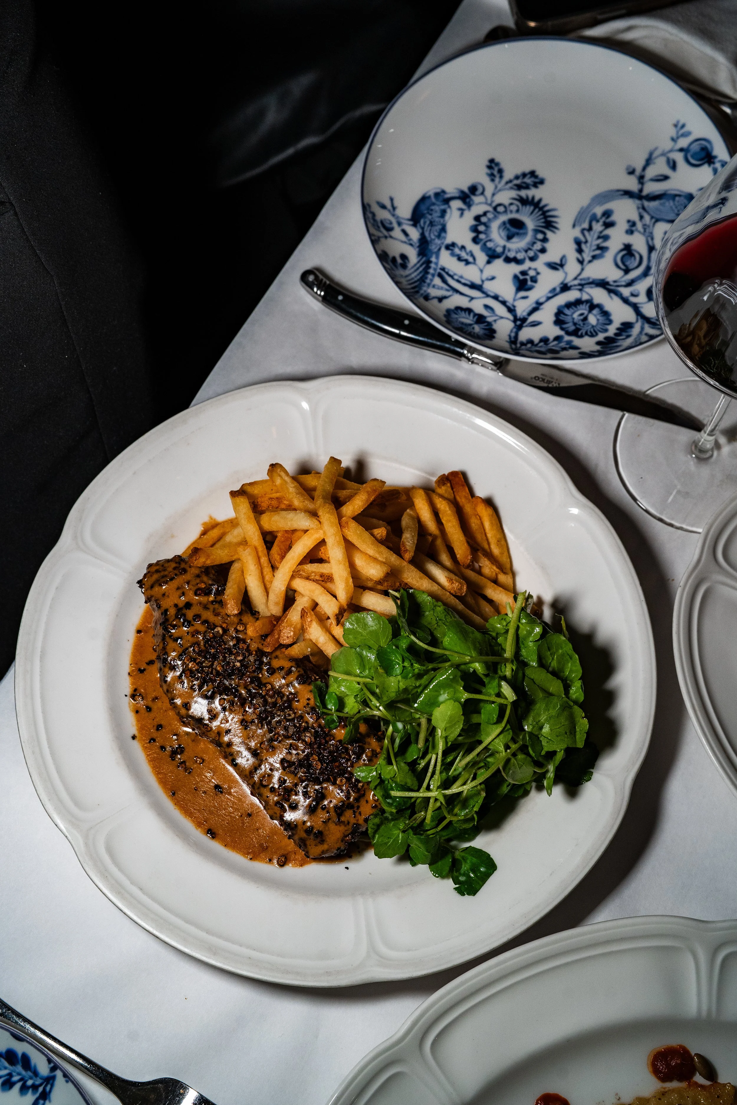 A plate with a cooked fish fillet topped with black pepper and a brown sauce, served with French fries and a side of fresh green watercress. On the table, there are blue and white patterned bowls, a glass of red wine, and a knife.