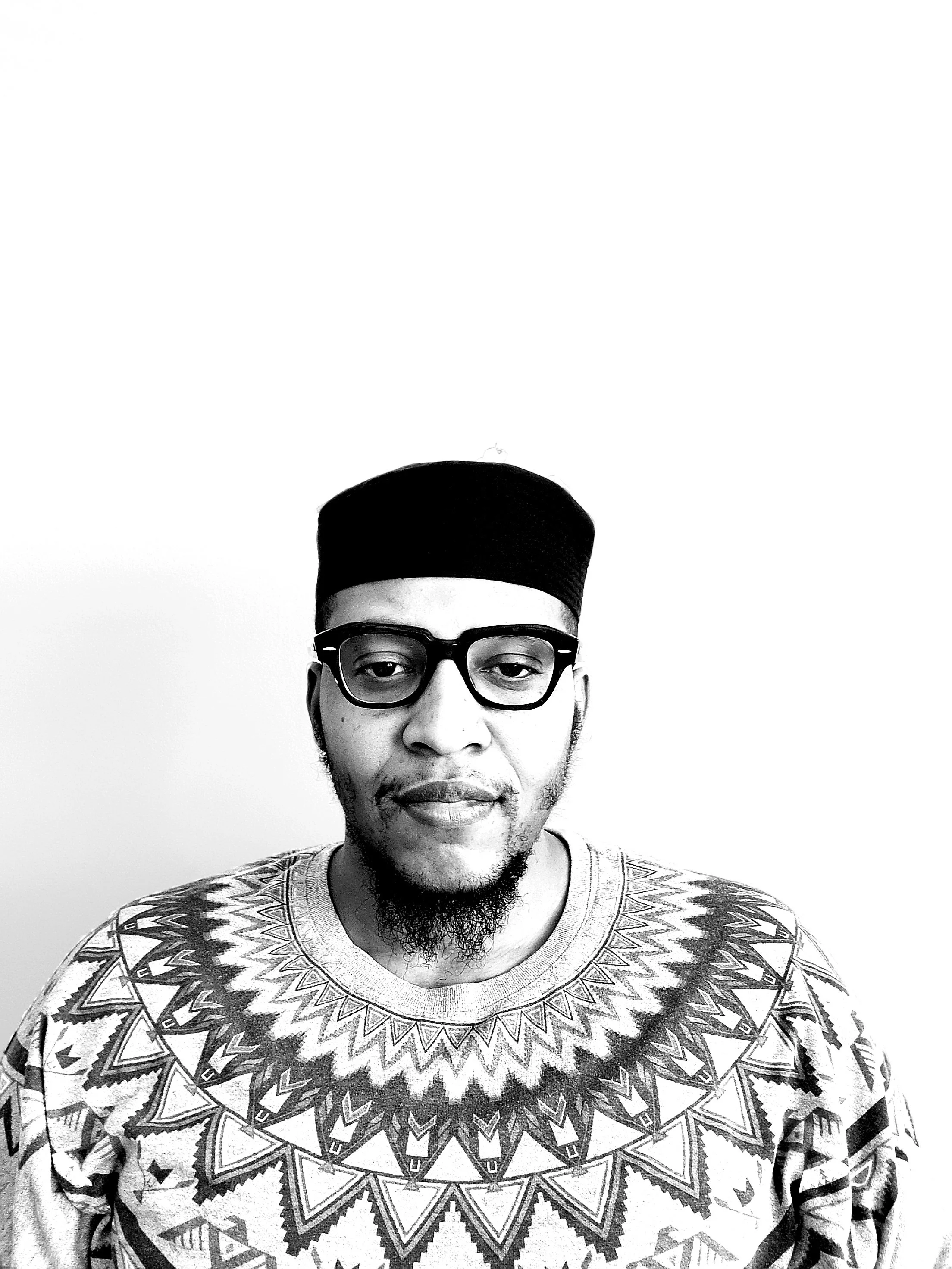 A black and white photo of a man wearing a brimless cap, thick frame Ray Ban glasses, and an Indigenous American inspired print t-shirt.