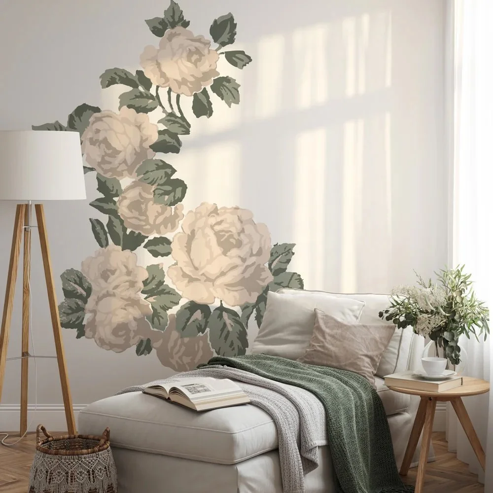 A cozy living room corner with a white chaise lounge, beige and green throw blankets, an open book, and pillows. There is a small wooden side table with a vase of white and green flowers, a white cup, and a book. A tall wooden floor lamp stands beside a floral wall mural of large, cream-colored roses with green leaves, and sunlight filters through sheer curtains.