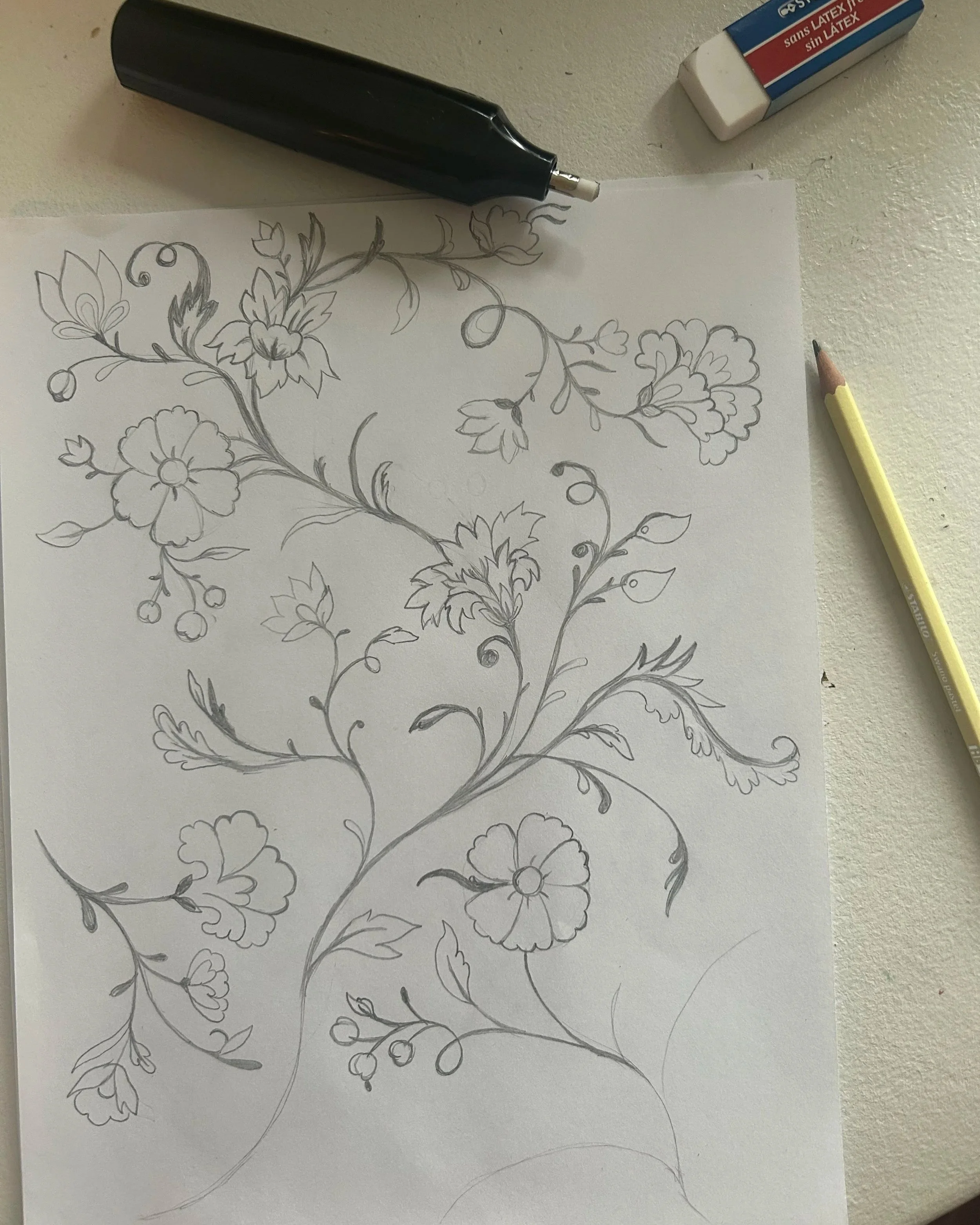 Sketch of a floral vine pattern with flowers, leaves, and small buds on a sheet of paper, with a pencil, an eraser, and a black tool nearby.