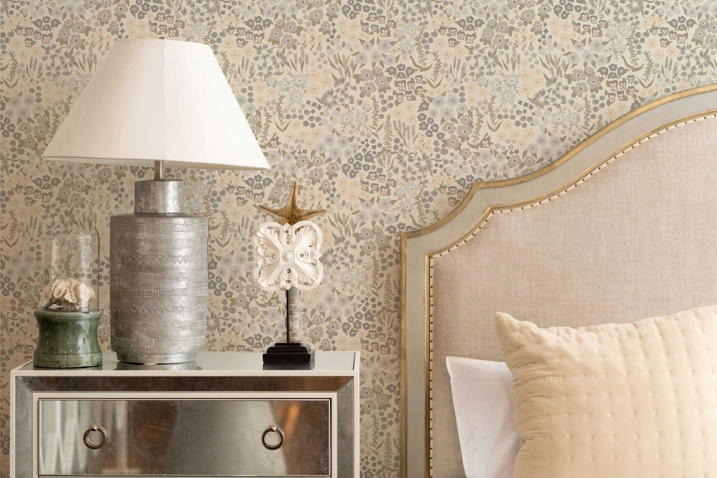 A bedroom nightstand with a large silver lamp, a small green pot with cream-colored flowers, and a decorative seashell sculpture with a starfish on top. Part of a beige upholstered bed with a large, textured pillow is visible.