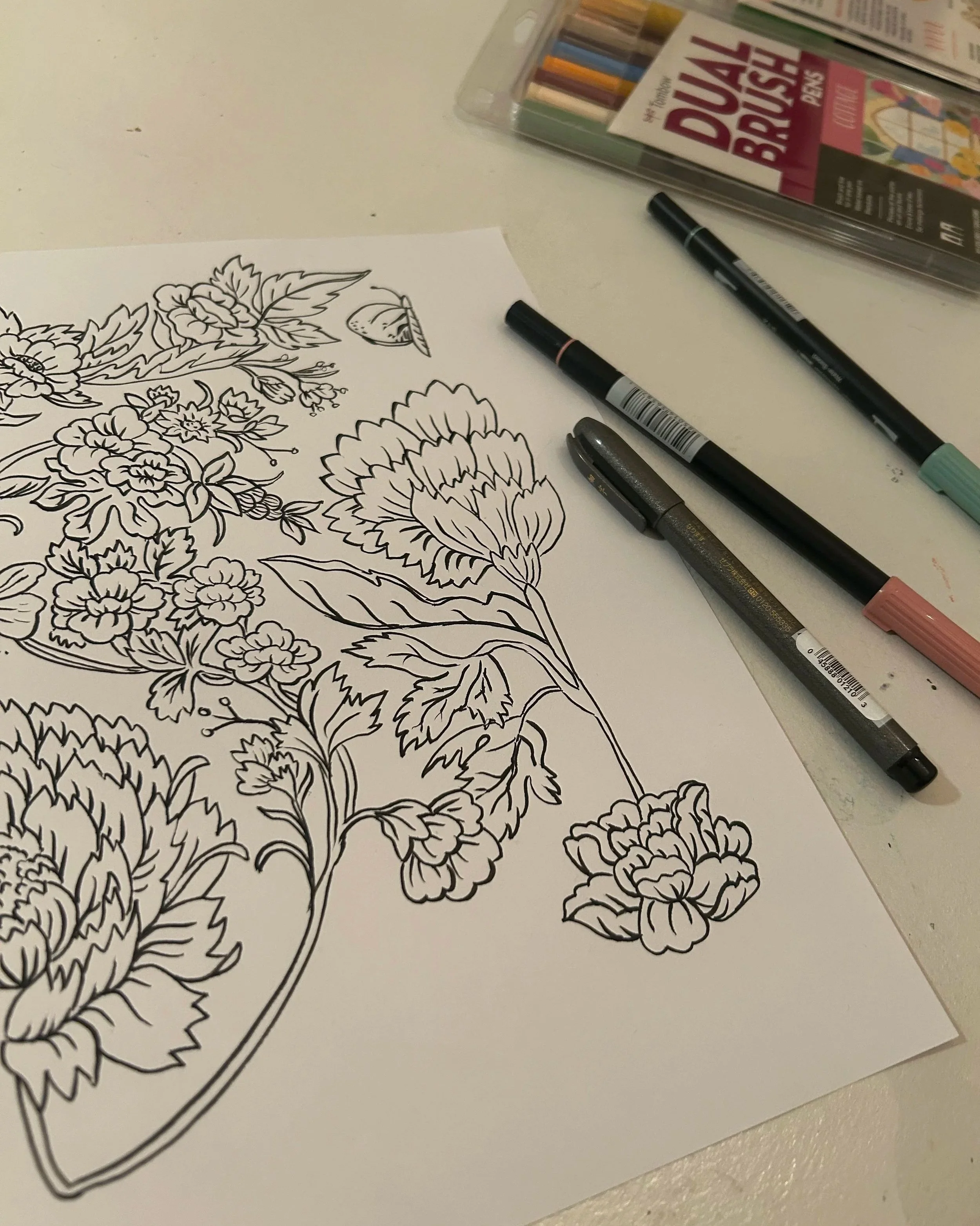 Line drawing of flowers on a sheet of paper placed on a white surface, with markers and a box of dual brush pens nearby.