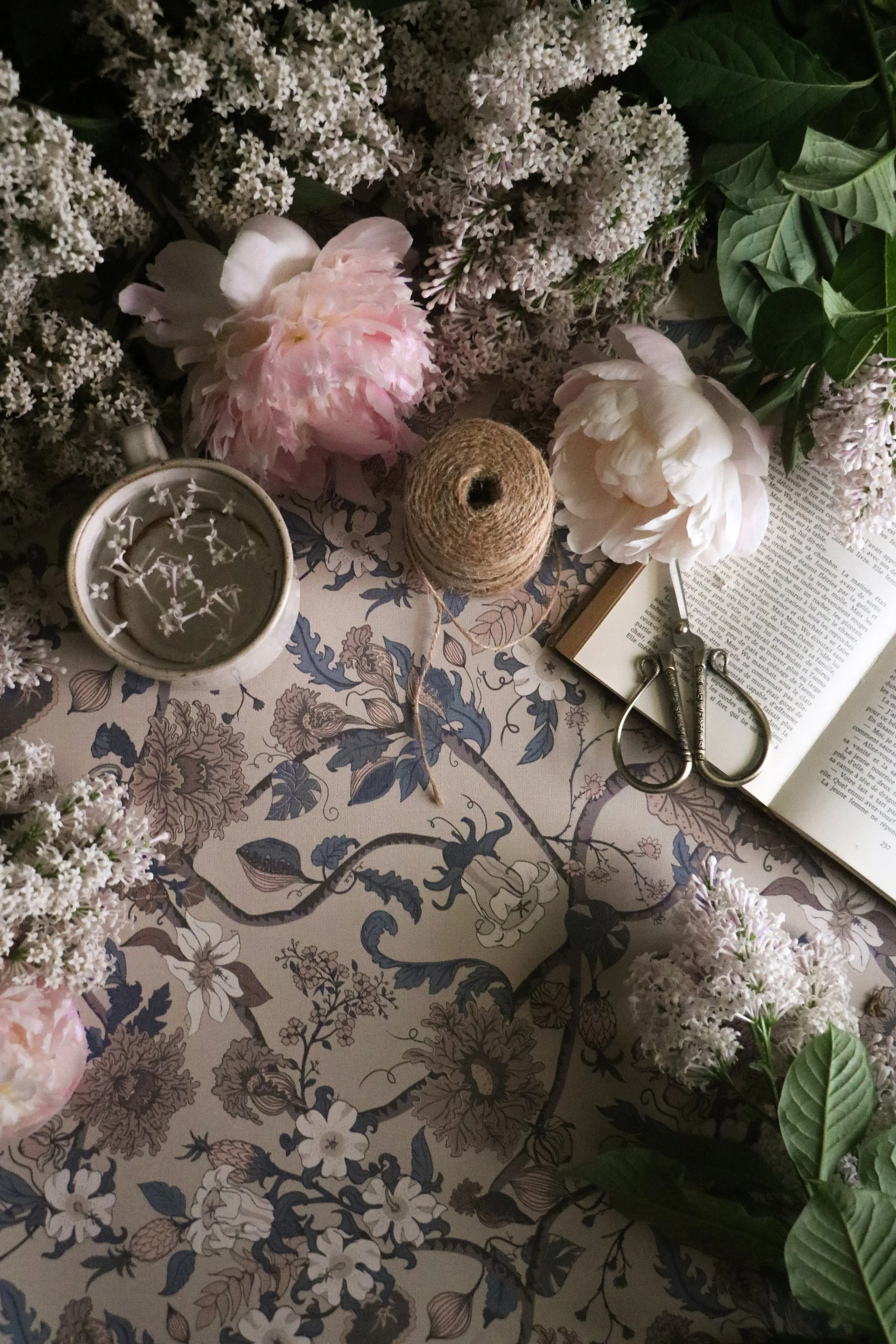 A floral wallpaper with pink and white peonies, floral wallpaper, a cup of tea with flower petals, a spool of twine, an open book, and scissors.