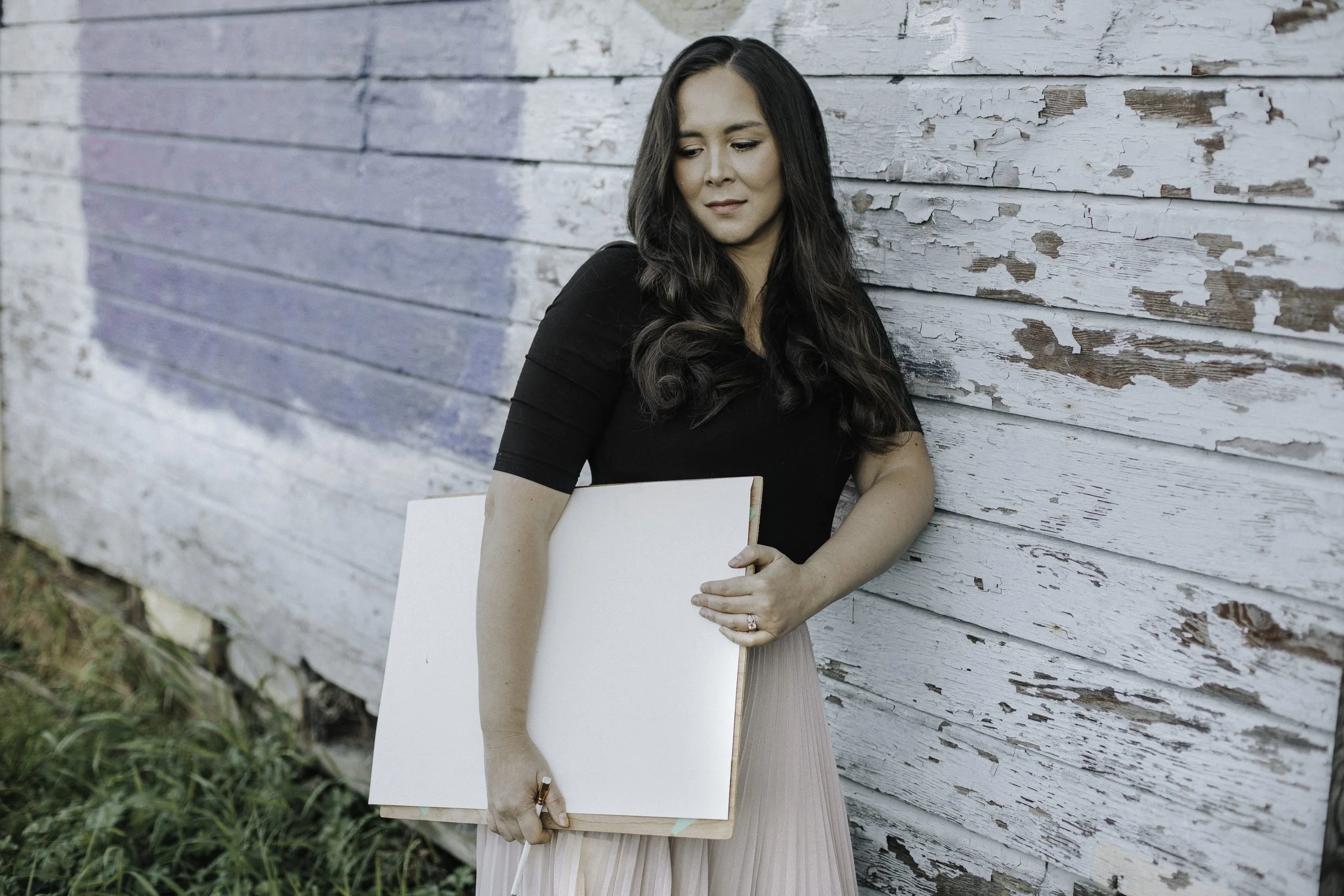 Gabrielle with long dark curly hair wearing a black top and pink pleated skirt leaning against a weathered white wooden wall, holding a blank canvas board.